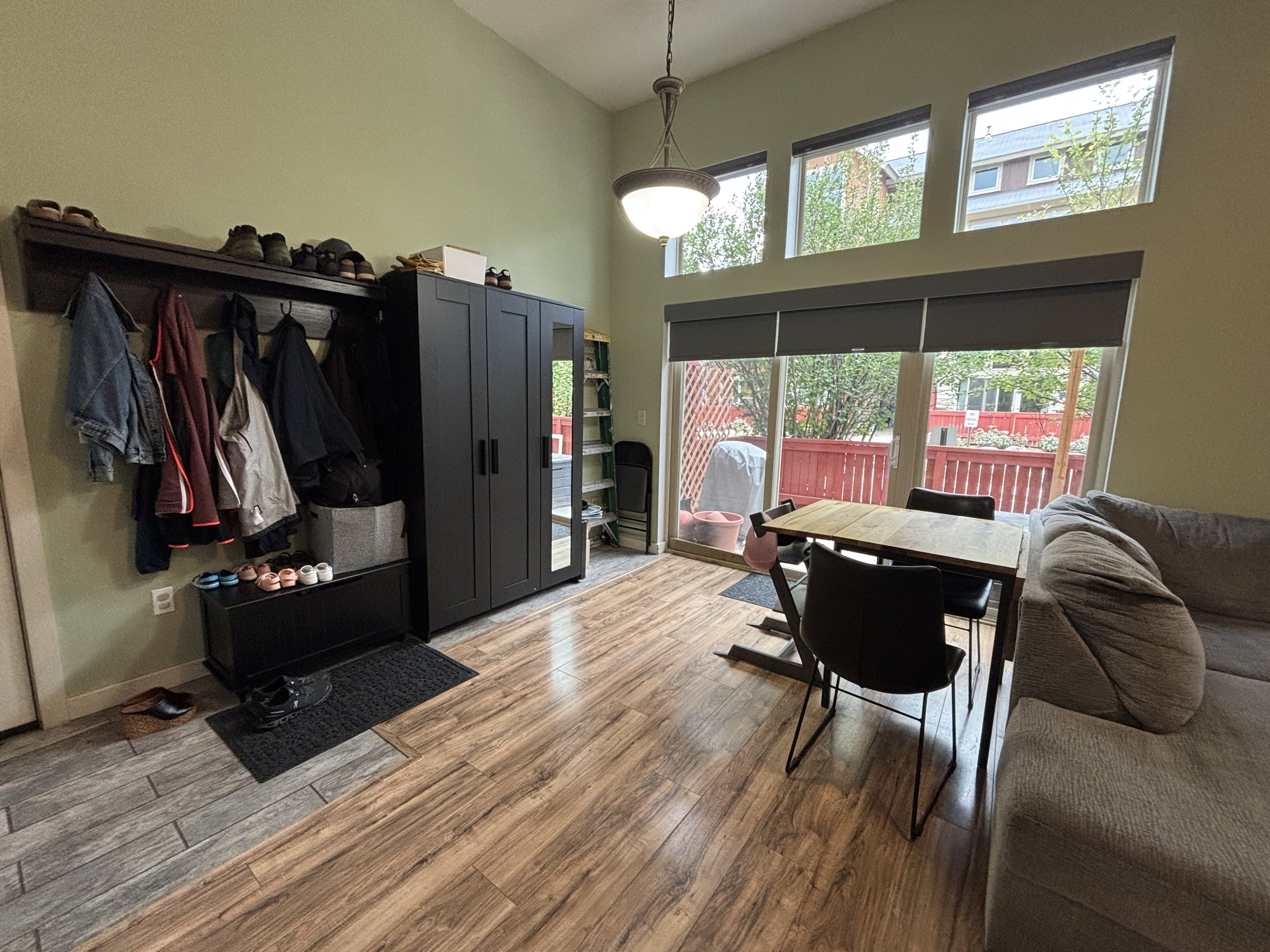 Interior view of a living room with a large window, a wooden dining table with black chairs, a gray sofa, a black storage unit with coats hanging above, shoes on a small black bench, and a sliding glass door leading to a backyard with a wooden fence 
