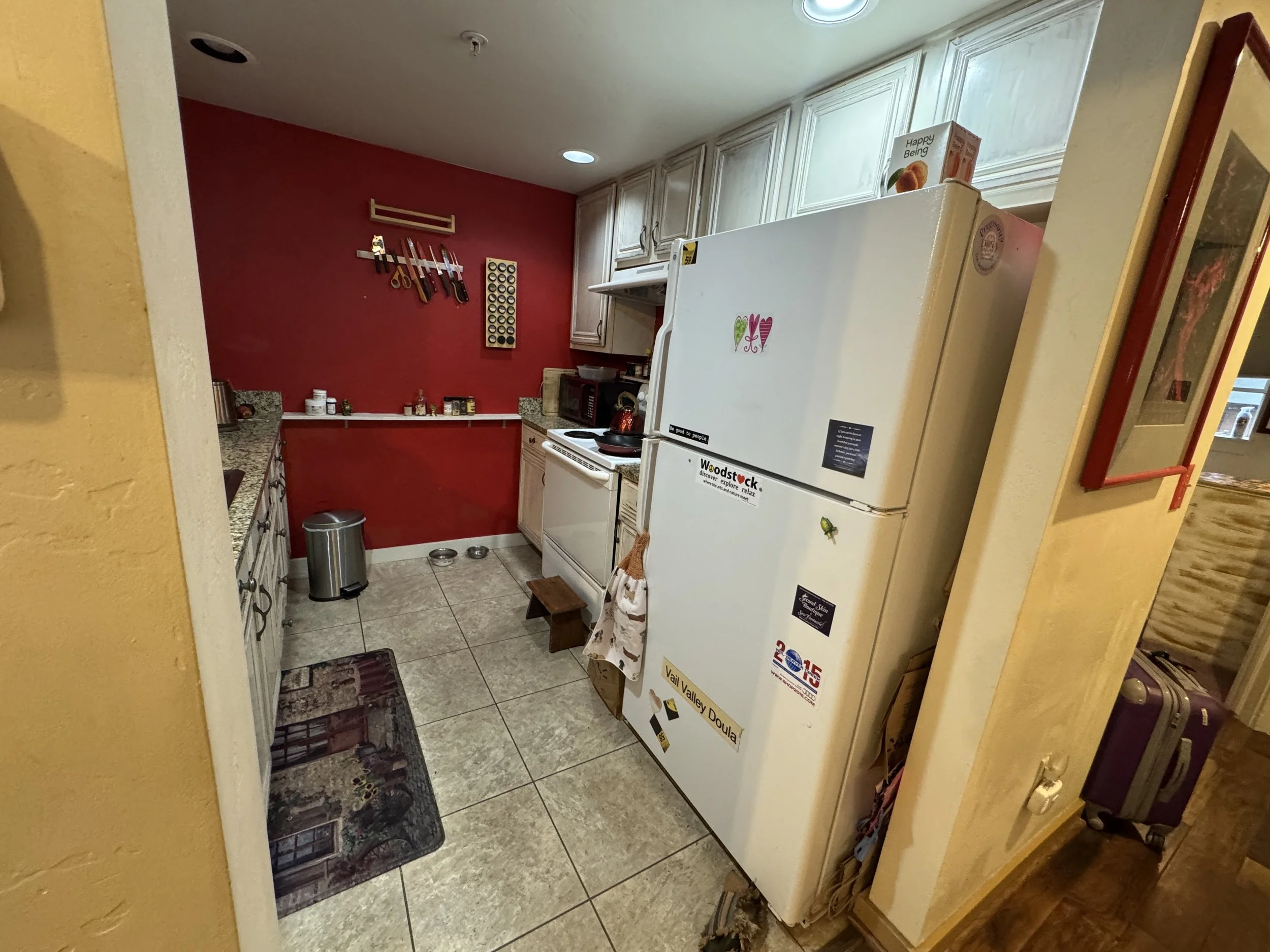 A kitchen with a red accent wall, white cabinetry, a white refrigerator with stickers, a stove, microwave, and various small items on the countertops. Knife rack and spice rack mounted on the red wall.