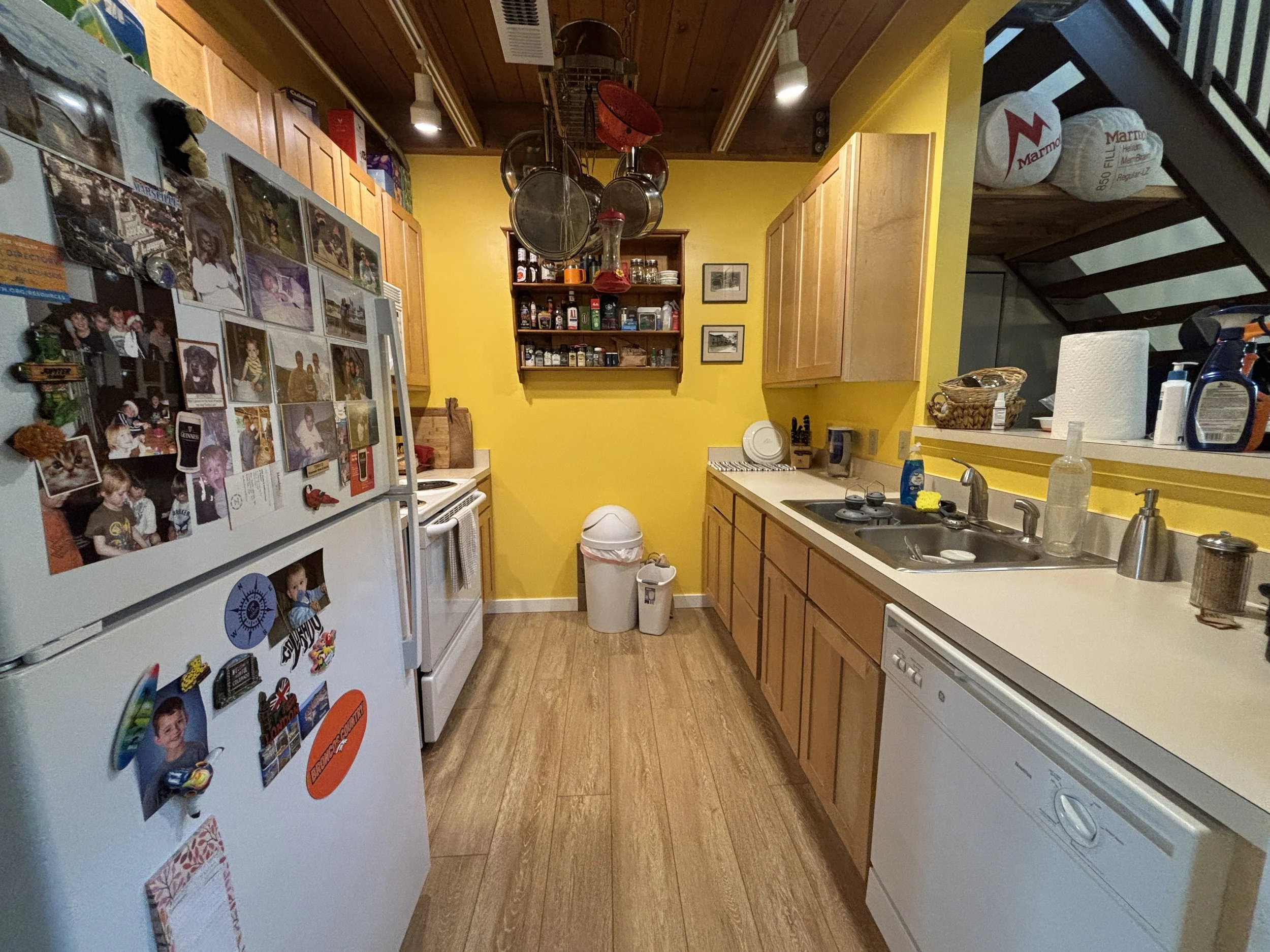 A kitchen with yellow walls, wooden cabinets, a white refrigerator covered with family photos and magnets, a countertop with cleaning supplies, a sink, and an overhead pot rack with pots and pans. There are two small framed pictures hanging on the wa