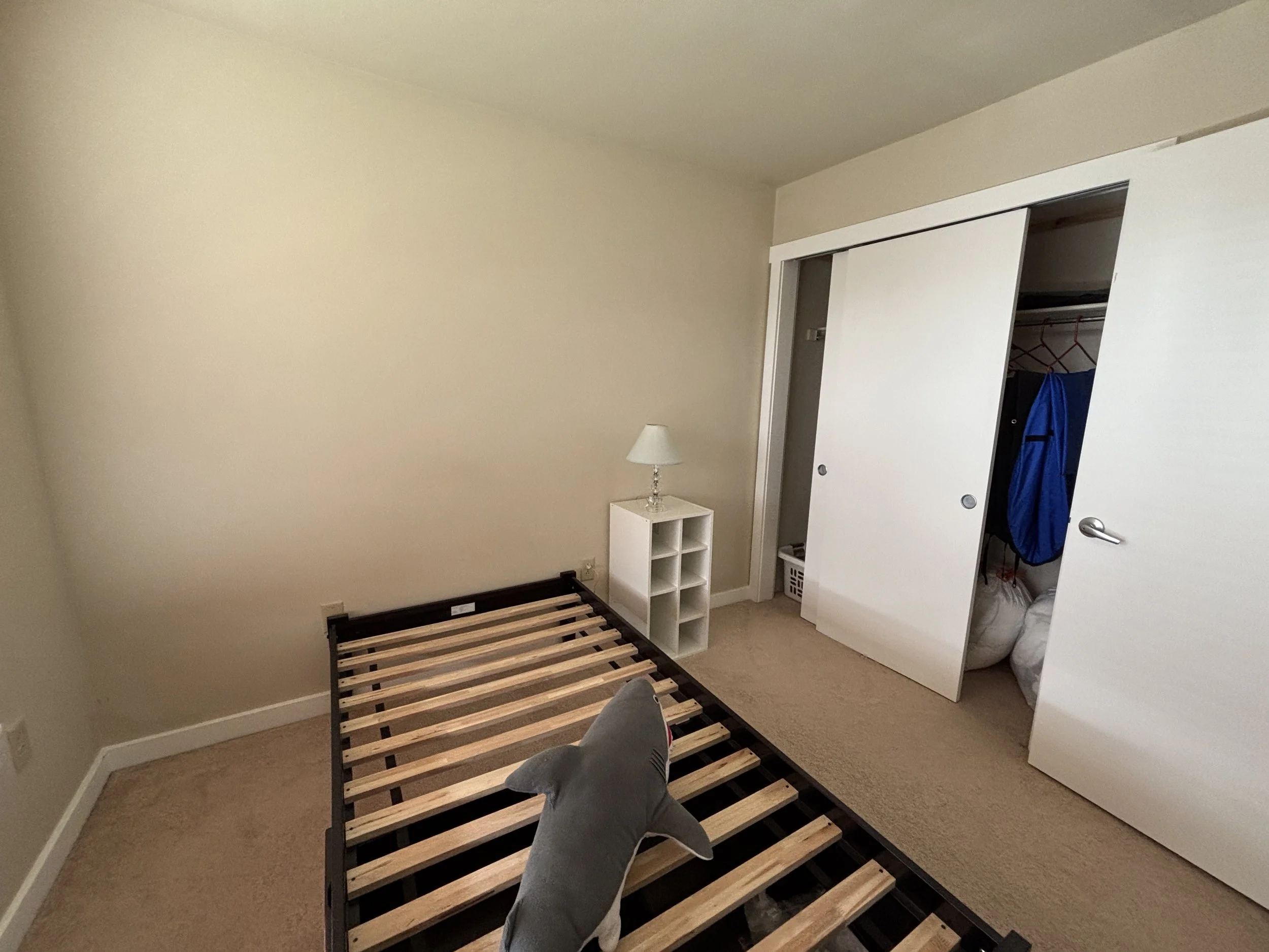 A bedroom with beige walls, a beige carpet, and white trim. There is an empty bed frame with wooden slats placed on a black metal frame. A small white bedside table with some white accessories is against the wall. To the right, there is a closet with