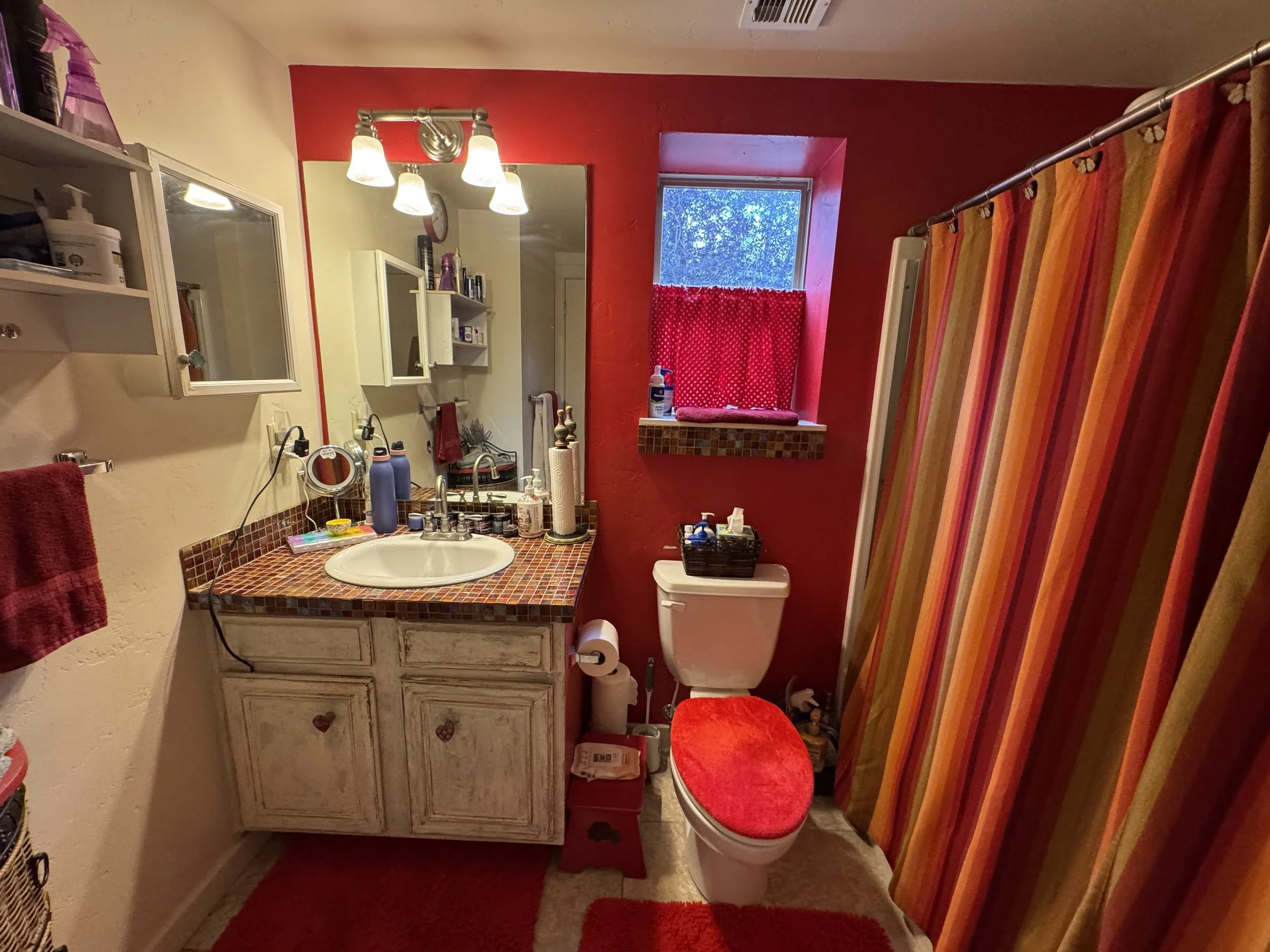 A small bathroom with red and cream walls, a white sink with a mosaic tile countertop, a mirror, and a window with a red polka dot curtain. There are various toiletries on the counter, a red towel, a red seat cover on the toilet, and a multi-colored 