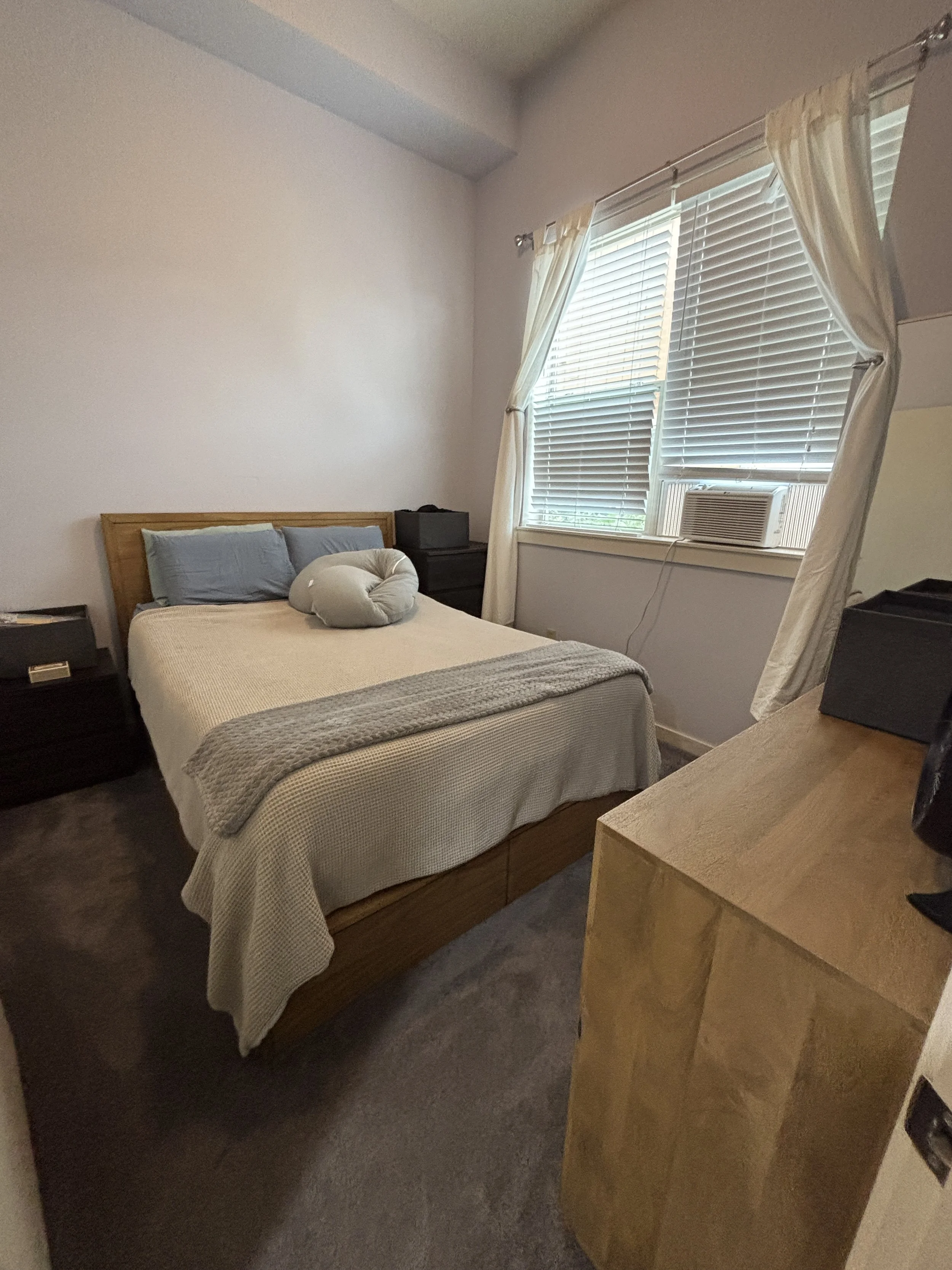 A bedroom with a bed that has a wooden frame, grey pillows, a white bedsheet, and a stuffed doughnut-shaped pillow. Next to the bed are two black nightstands, and a window with white blinds, partially open, with cream-colored curtains. An air conditi
