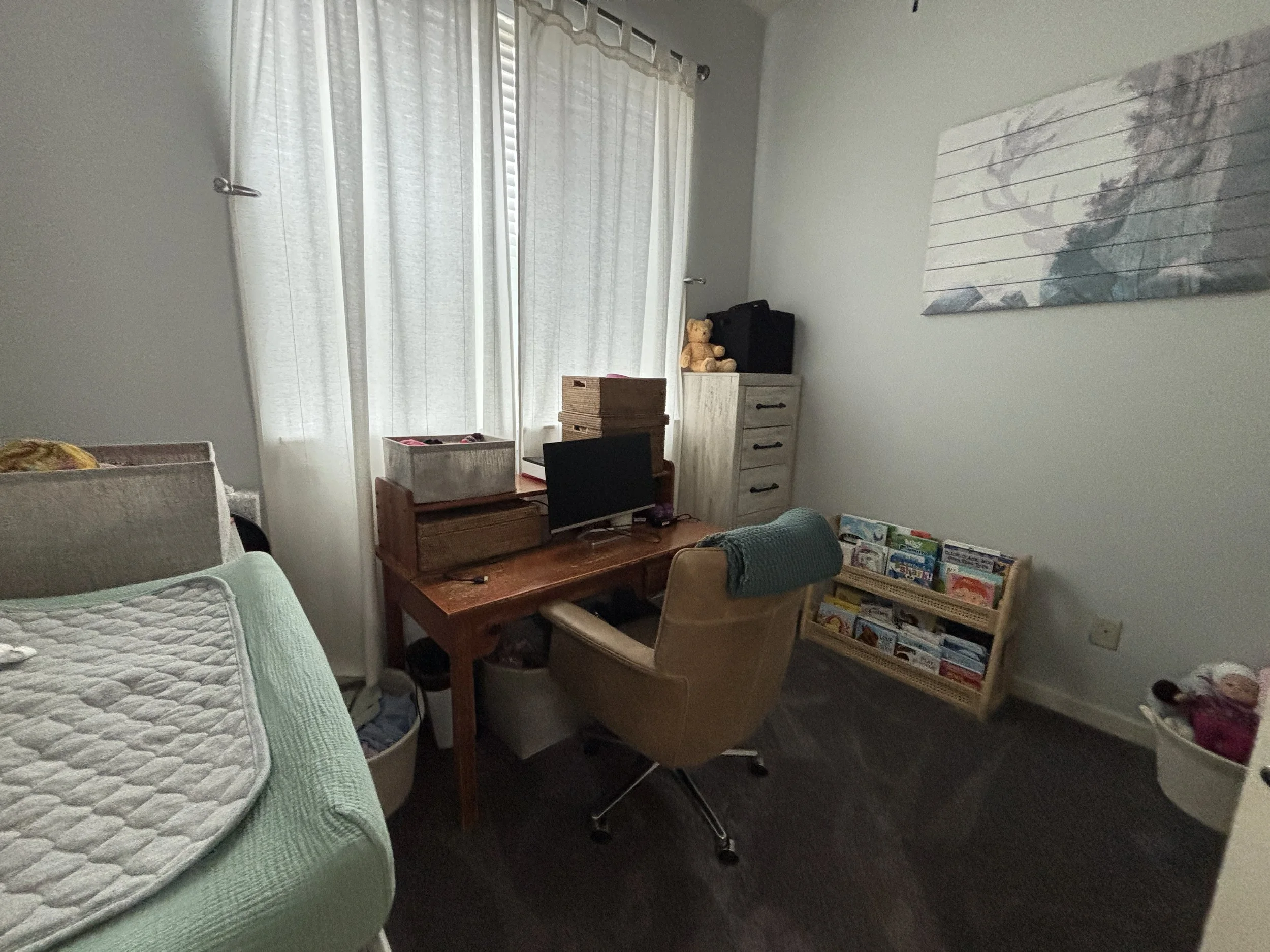 A room with a bed on the left side with a green blanket and a quilted mattress. In the middle, there is a wooden desk with a chair, some storage boxes, and a small TV. To the right, there is a beige armchair with a teal blanket draped over it and a b