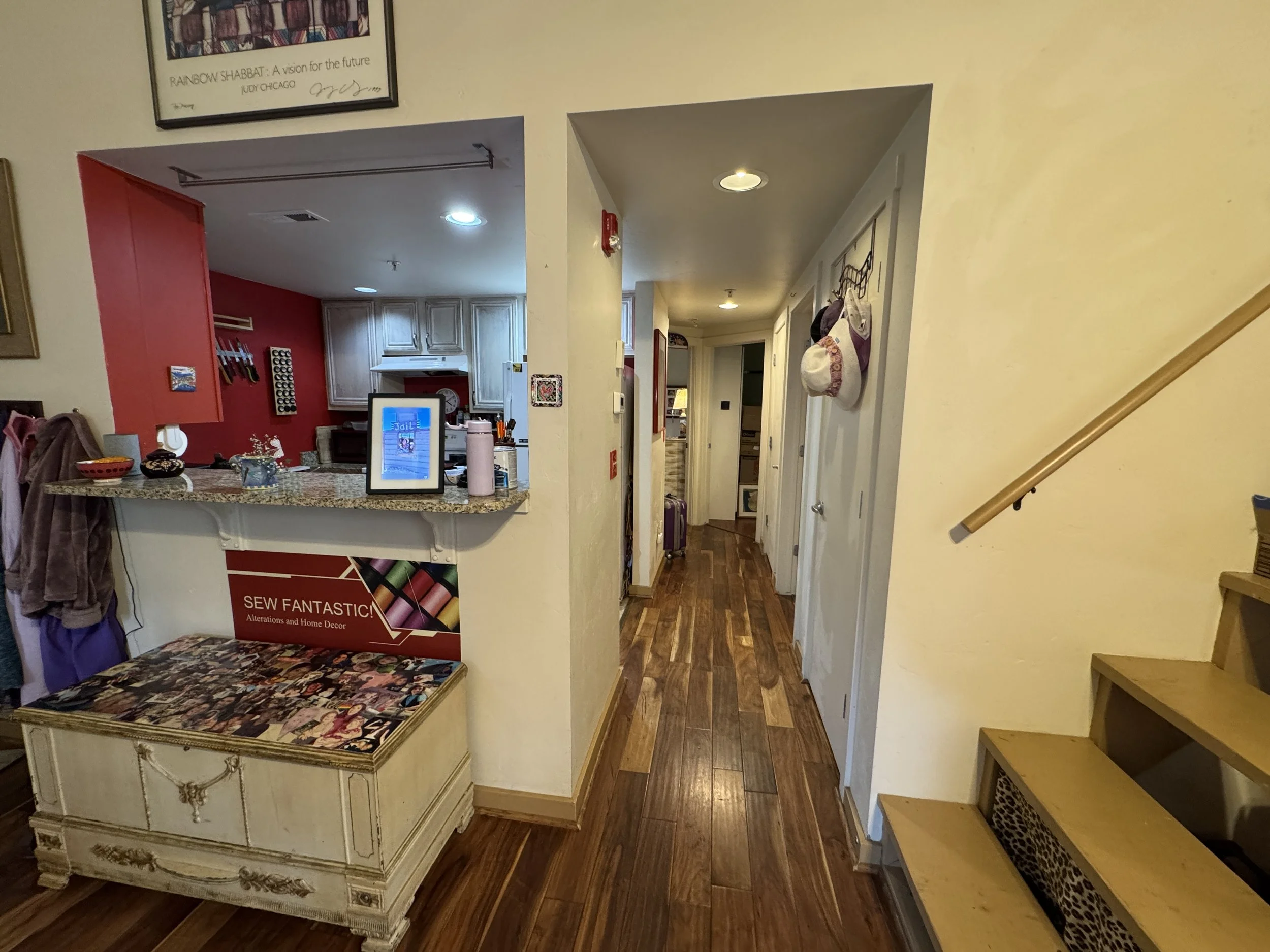 The image shows a hallway with wooden flooring. To the left, there is a partially visible white carved wooden dresser with a collage of images on its top surface. The hallway leads to a kitchen area with white cabinets and a red accent wall with kitc