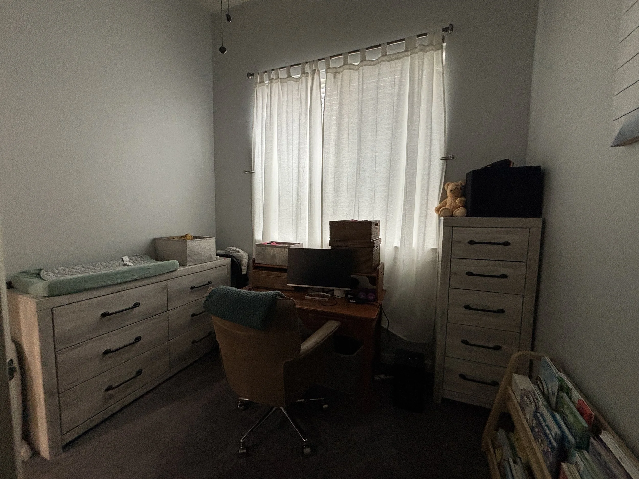 A dimly lit bedroom with a bed, dresser, and bookshelf, a window with curtains, and various objects like a teddy bear and books.