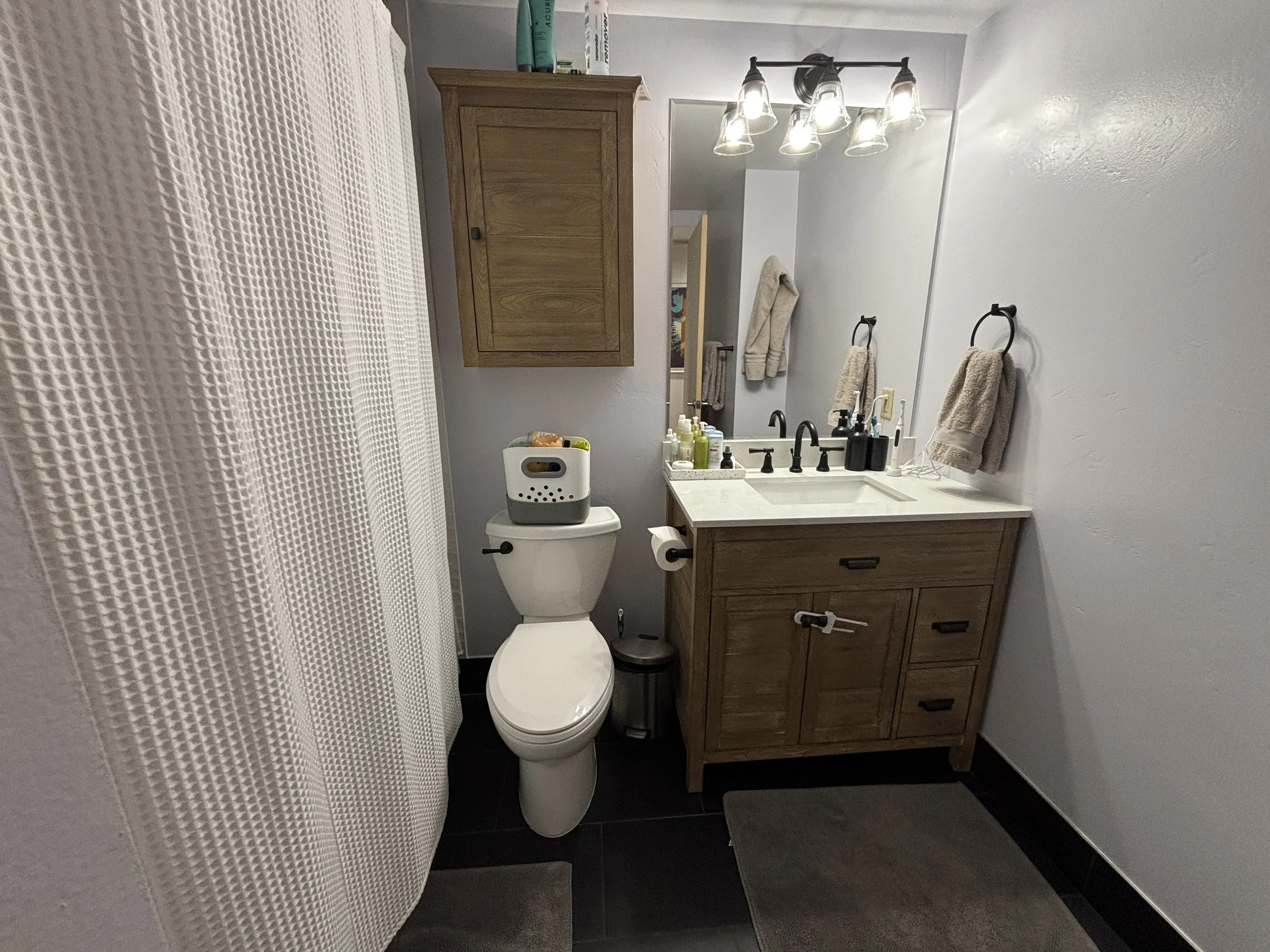 A bathroom with a toilet, wooden vanity with a sink, mirror, and light fixture with multiple bulbs. There are toiletries on the vanity and a towel hanging on the wall. The floor is dark-colored tile, and a white curtain is partially visible on the le