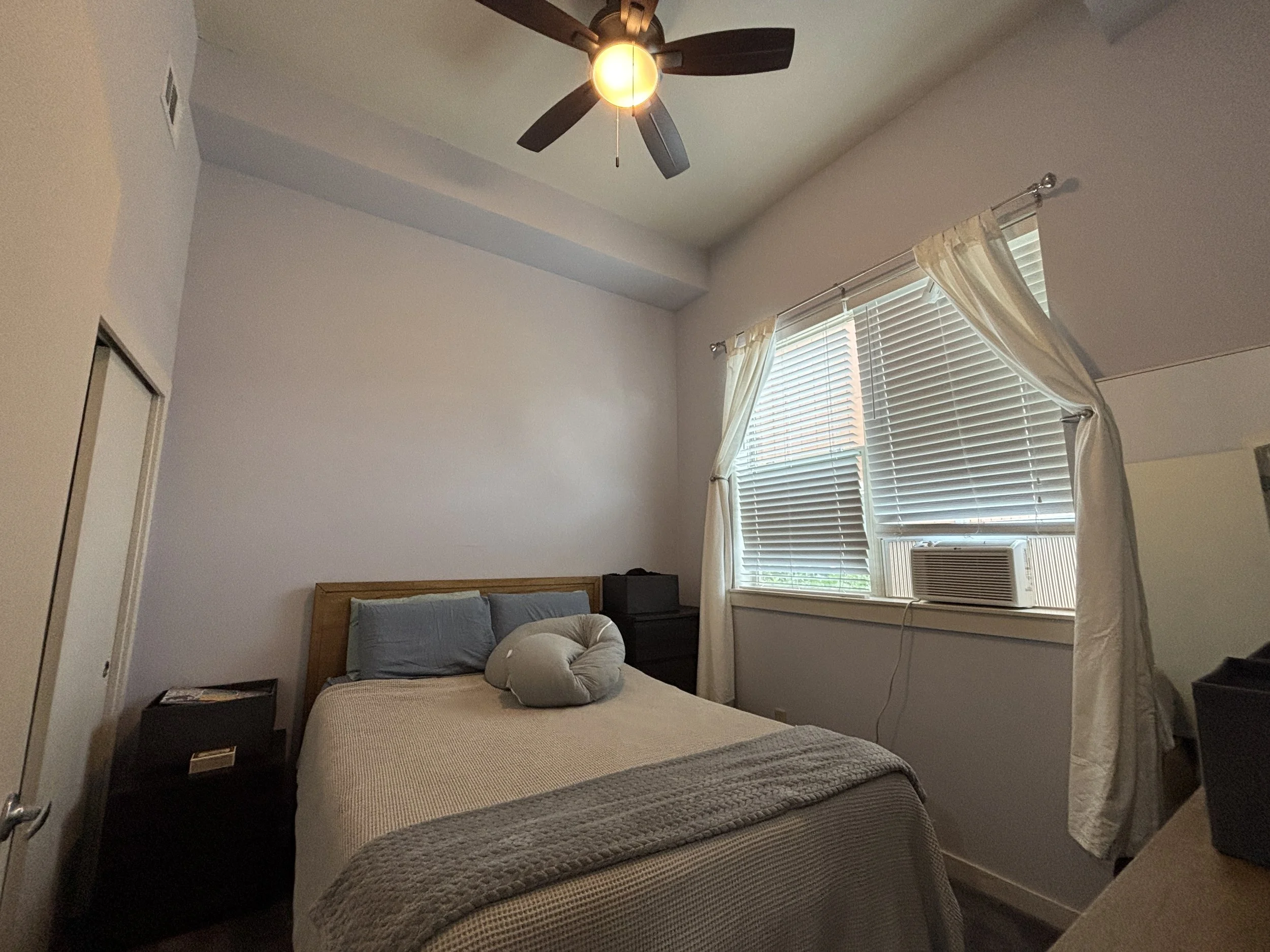 A bedroom with a wooden bed, gray bedding, decorative pillow, bedside table, window with white blinds and curtains, ceiling fan with light.