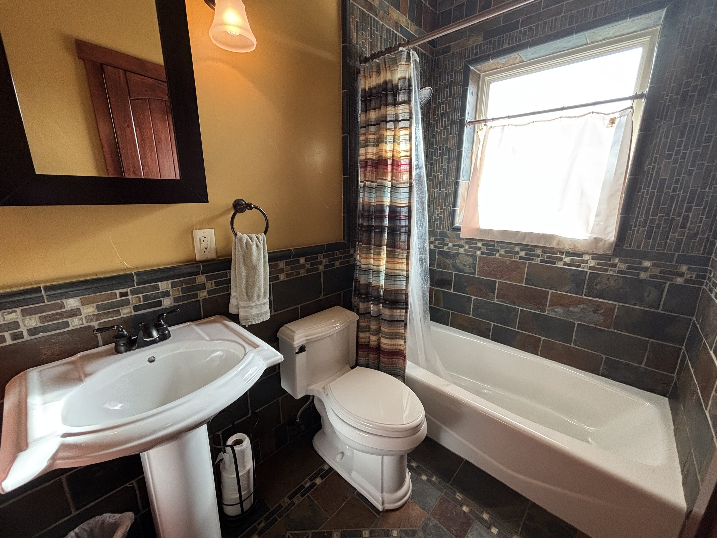 Bathroom with brick tile walls, a bathtub with a shower curtain, a toilet, a pedestal sink, a mirror, a towel ring with a towel, and a window covered with a small curtain.