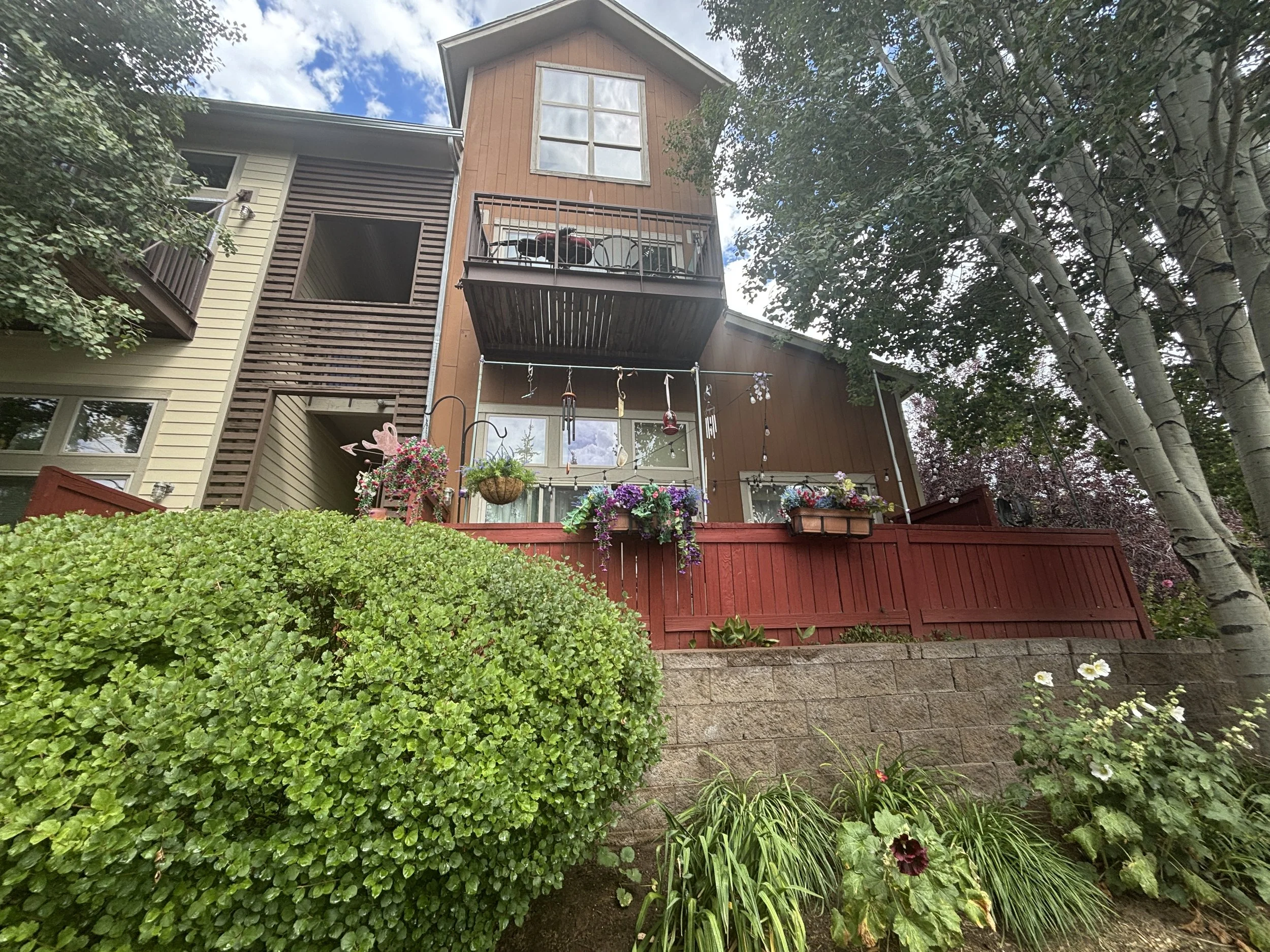 A multi-story house with a mixture of beige and brown siding, large windows, and a small balcony. The house is surrounded by lush green trees and shrubs, with flower pots and hanging decorations in the foreground.