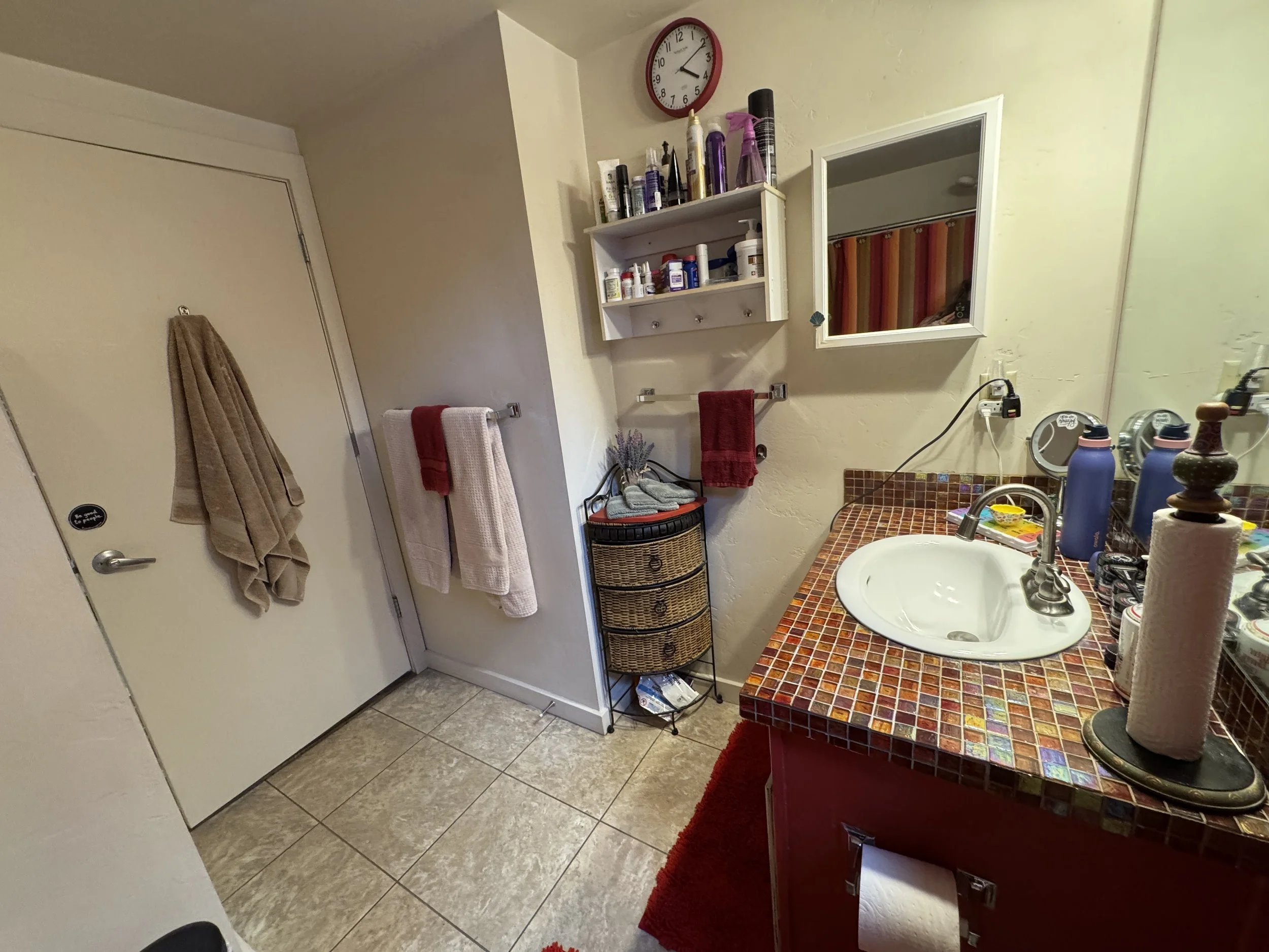 A bathroom with a white door, brown and white towels hanging on a wall hook, a small white shelf with various toiletries, a mirror and sink with a colorful tiled countertop, and a wicker laundry basket in the corner.