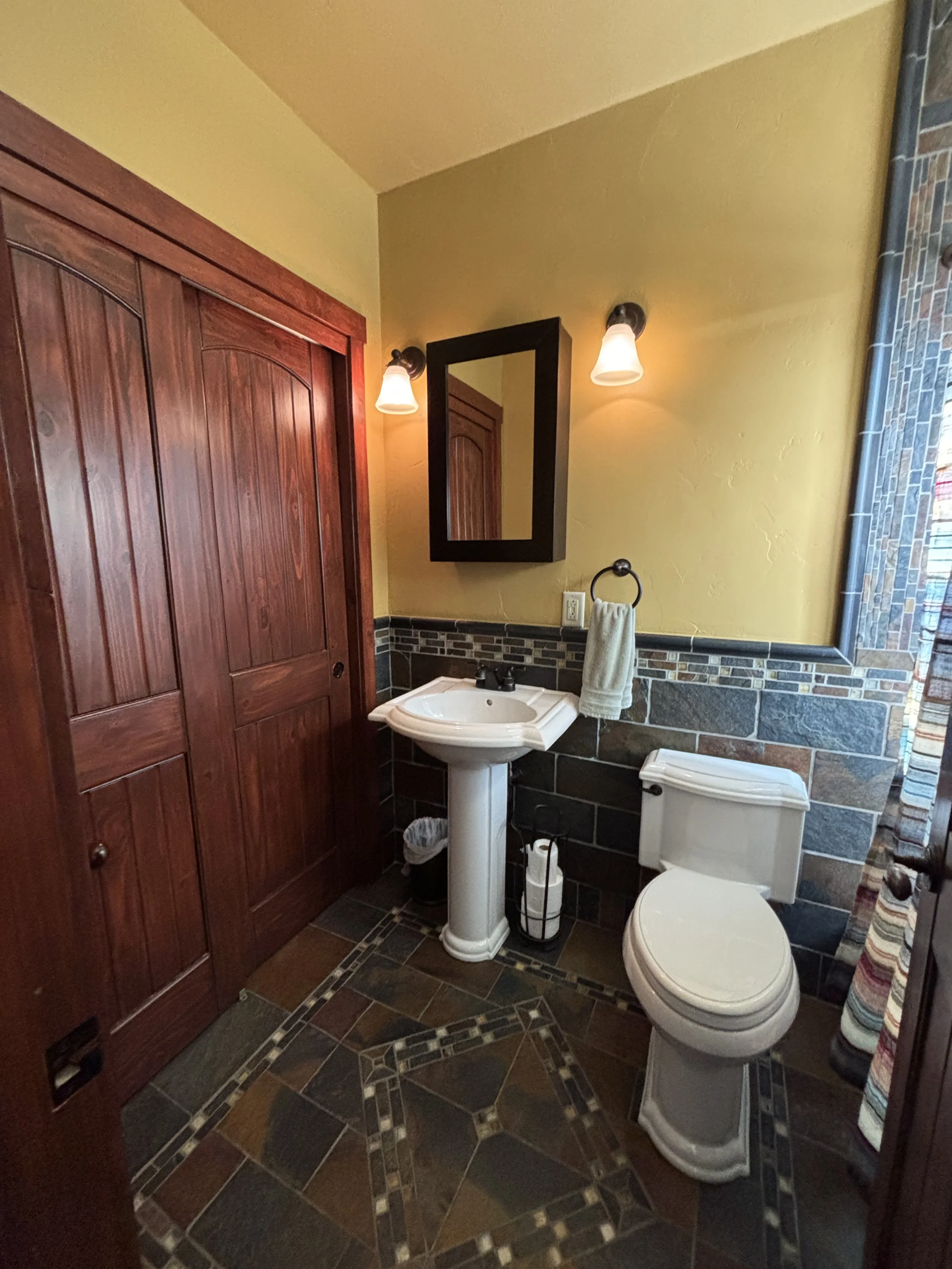 A bathroom with yellow walls and a decorative tile border. It features a wooden closet, a pedestal sink with a mirror above, and a toilet. Two wall-mounted lights illuminate the space, and a towel hangs on a ring near the sink.