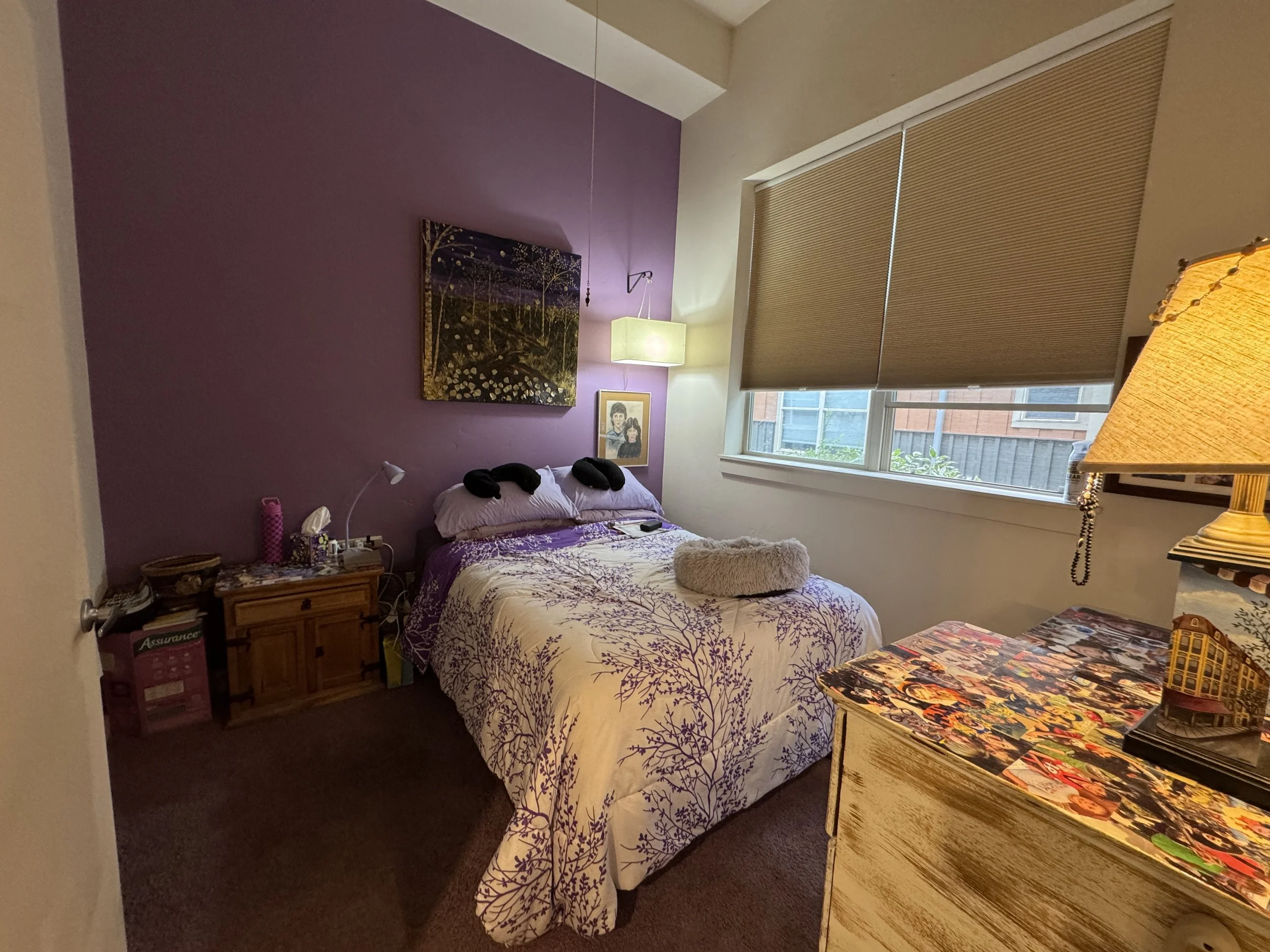 A cozy bedroom with purple and beige walls. There is a bed with white and purple bedding and black pillows. A small framed picture hangs above the bed. A wooden nightstand with various items, including a pink water bottle, is beside the bed. A large 