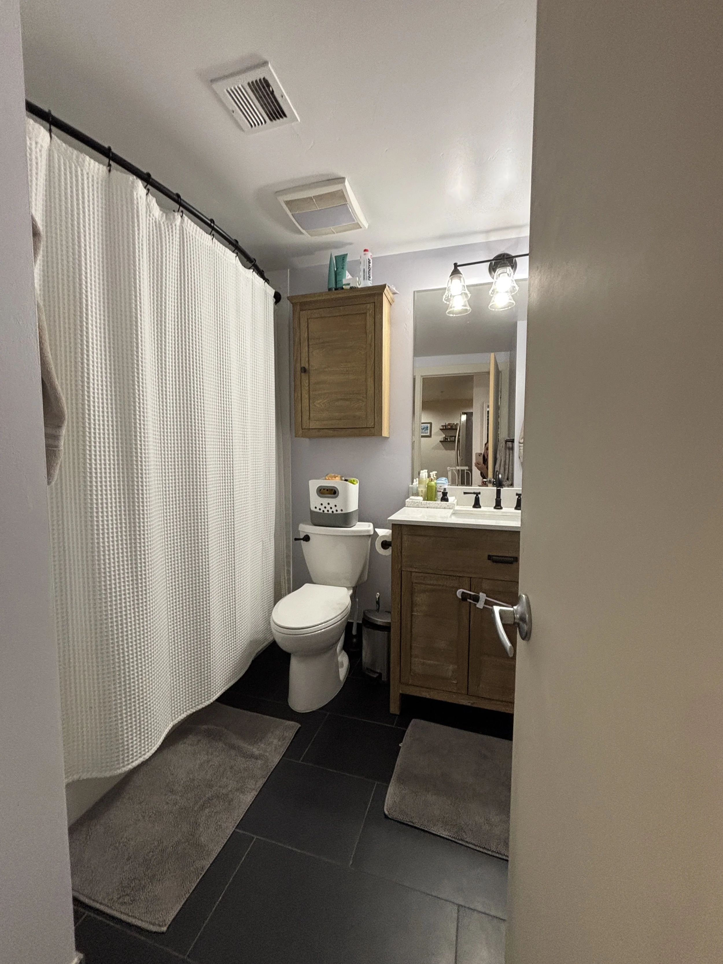 Bathroom with a toilet, wooden vanity cabinet with a sink, mirror, and wall cabinet, shower with white curtain, items on the sink, and a gray bath mat on the black tiled floor.