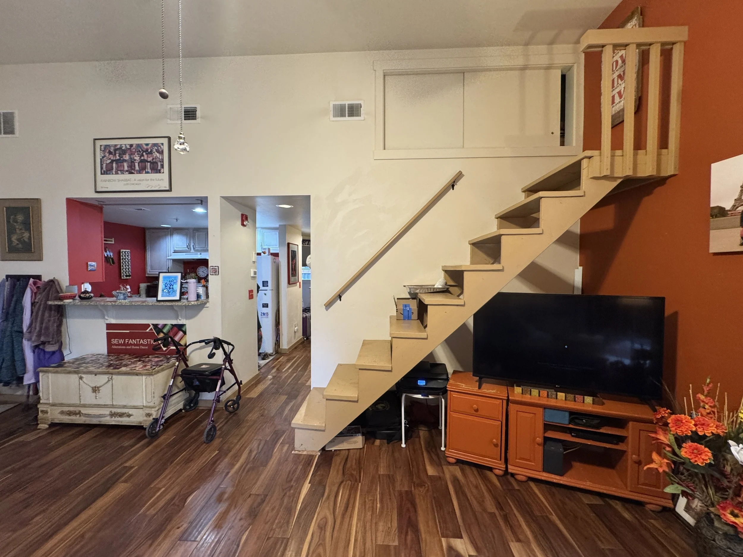 A living room with wooden flooring, a staircase on the right, a television, and various furniture and decorations, including a table with flowers, a framed artwork on the wall, and a kitchen area in the background.