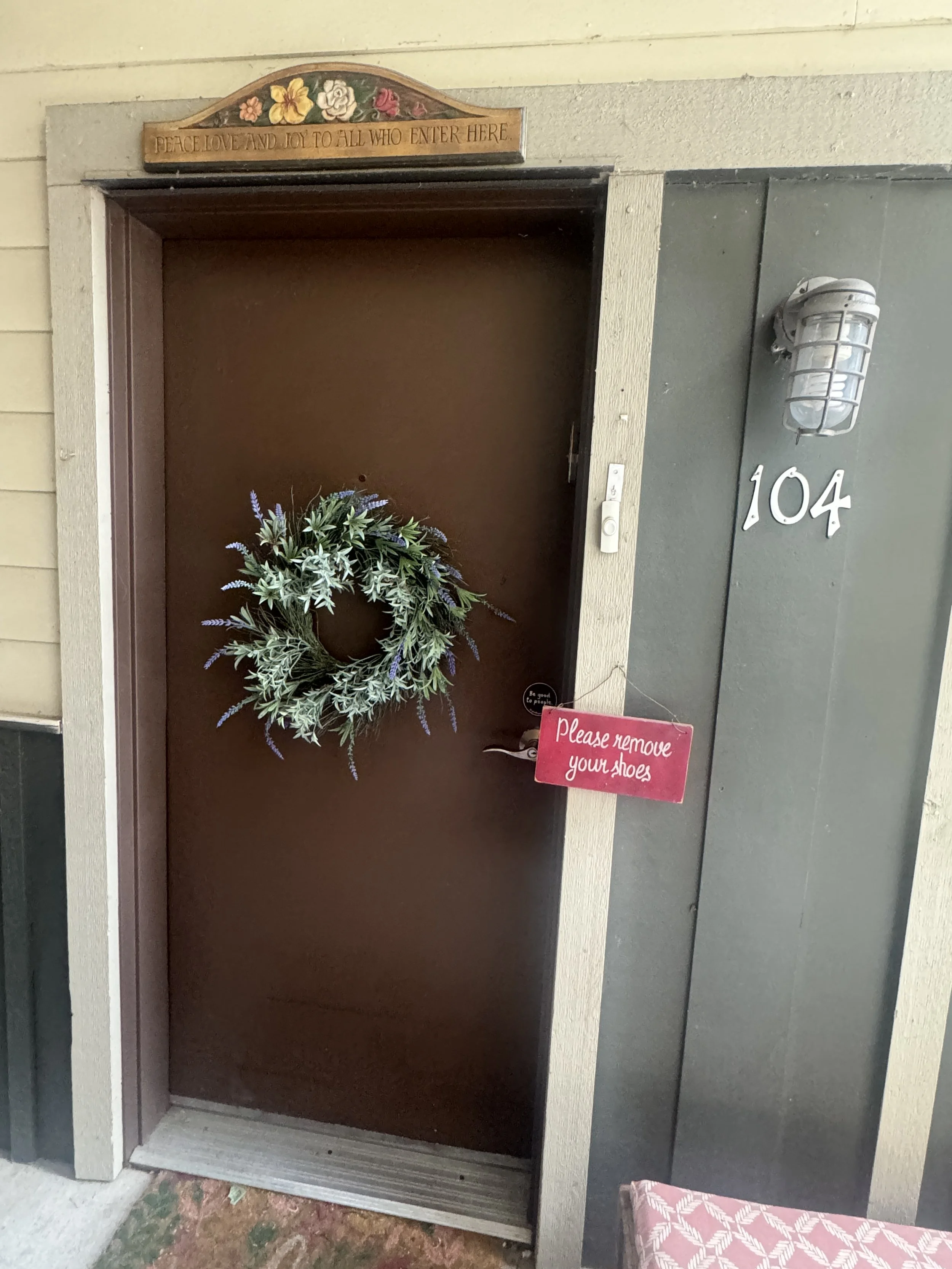 Front porch of a house with a brown door, a floral wreath hanging on it, a sign above the door reading "PEACE, LOVE AND JOY TO ALL WHO ENTER HERE," and a small pink sign on the door handle that says "Please remove your shoes." There is a gray wall wi