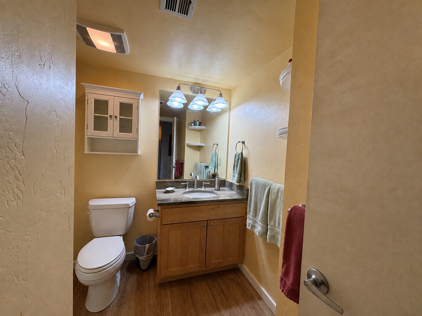 A small bathroom with yellow walls, a mirror above a wooden vanity with a sink, a toilet, and a small cabinet on the wall. Towels hang on rings, and a ceiling light fixture with four bulbs illuminates the space.