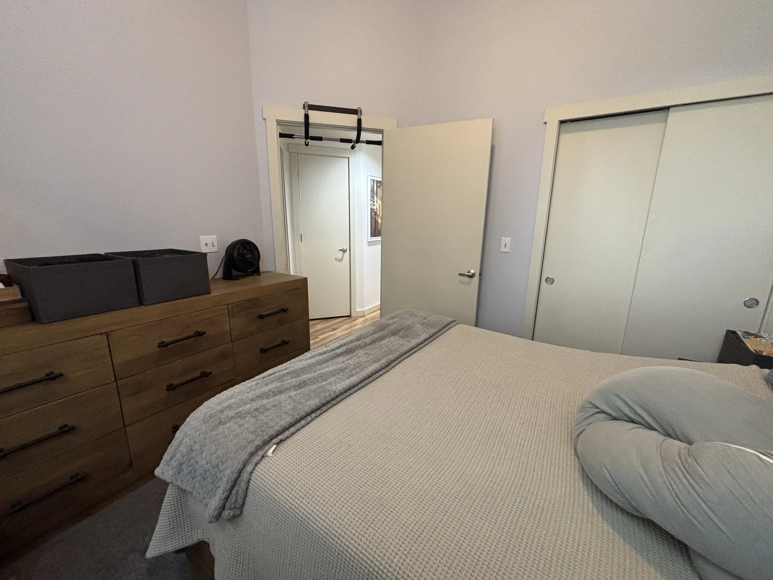 A bedroom with a bed, dresser, closet, and door. The bed has a gray blanket and pillow, and the dresser has three black boxes and a small fan. The closet has sliding white doors, and the door is open. The walls are painted light gray.