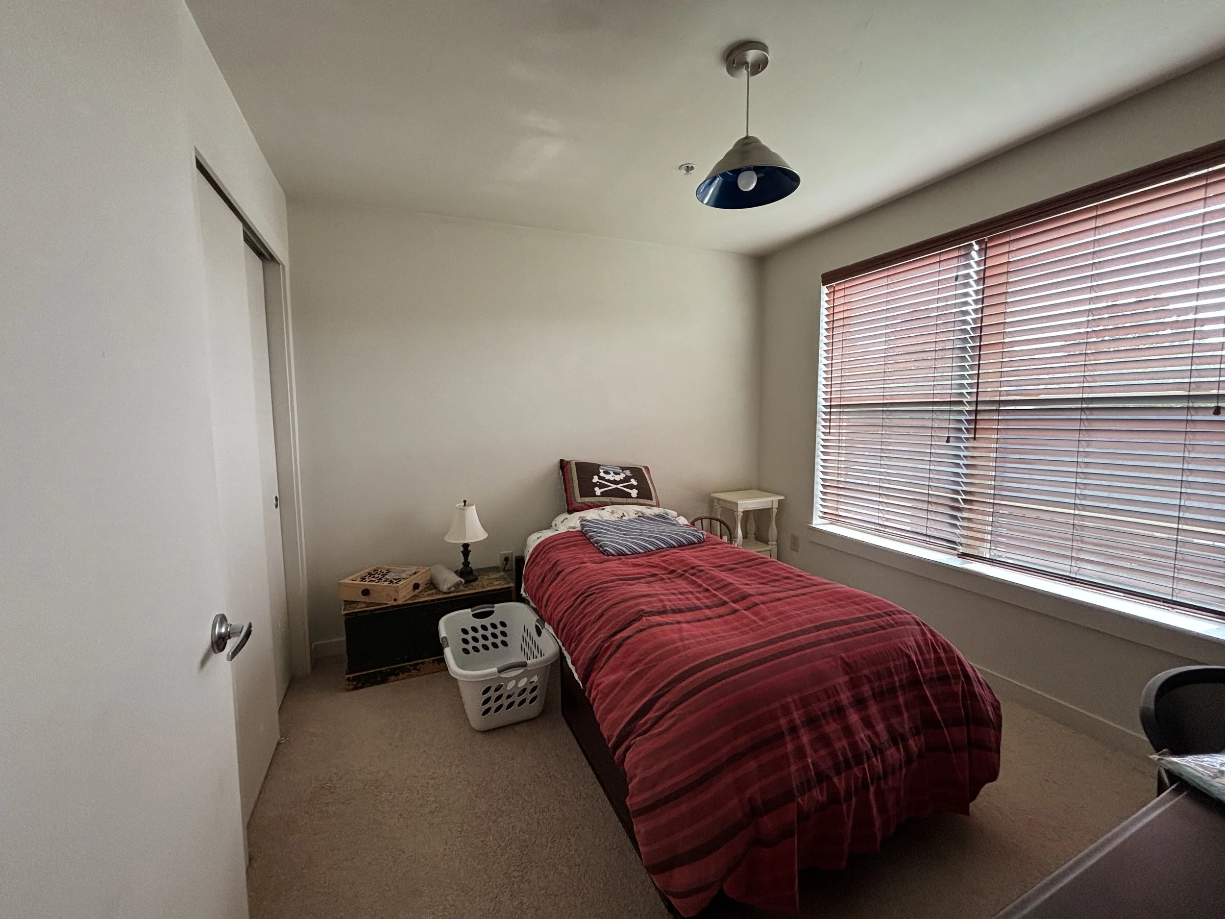 A small bedroom with a twin bed covered in red and black plaid bedding, a white pillow, and a pirate-themed pillow. There is a small nightstand with a lamp and a crate at the foot of the bed. The room has a large window with horizontal blinds, a beig