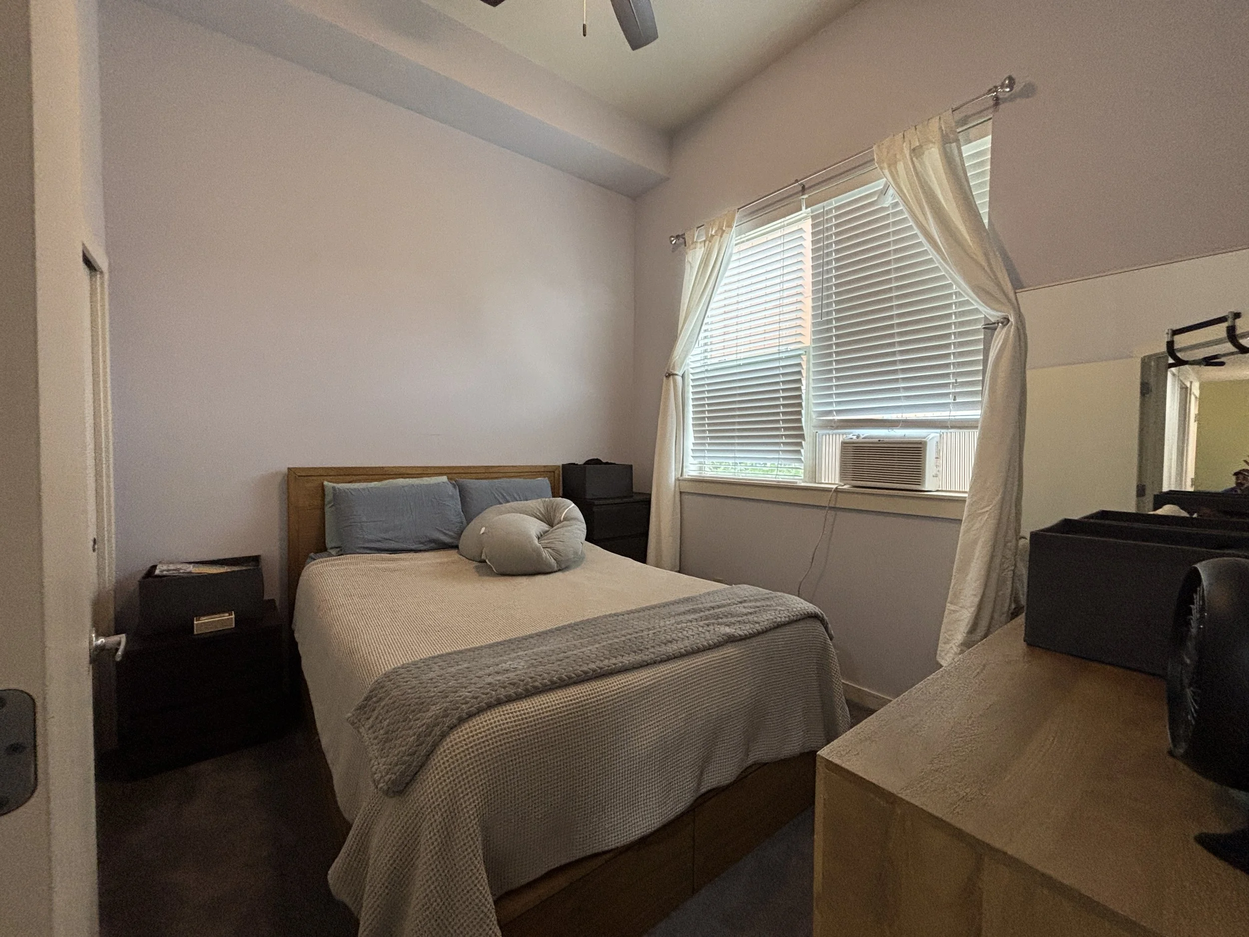 A bedroom with a wooden bed frame, a beige bedspread, and two pillows. There is a nightstand with a book and a small black speaker. The room has a window with white blinds and cream-colored curtains, and an air conditioning unit below the window. A c