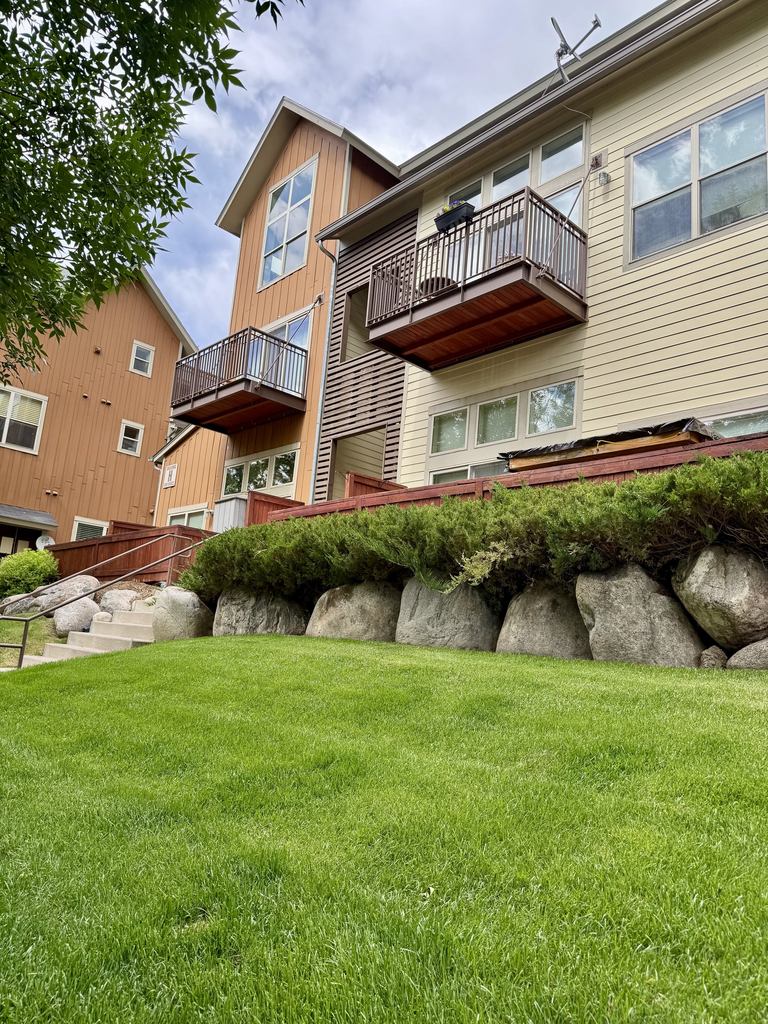 Front view of multi-story residential buildings with balconies, beige and brown siding, surrounded by a well-maintained green lawn, large rocks, and shrubs.