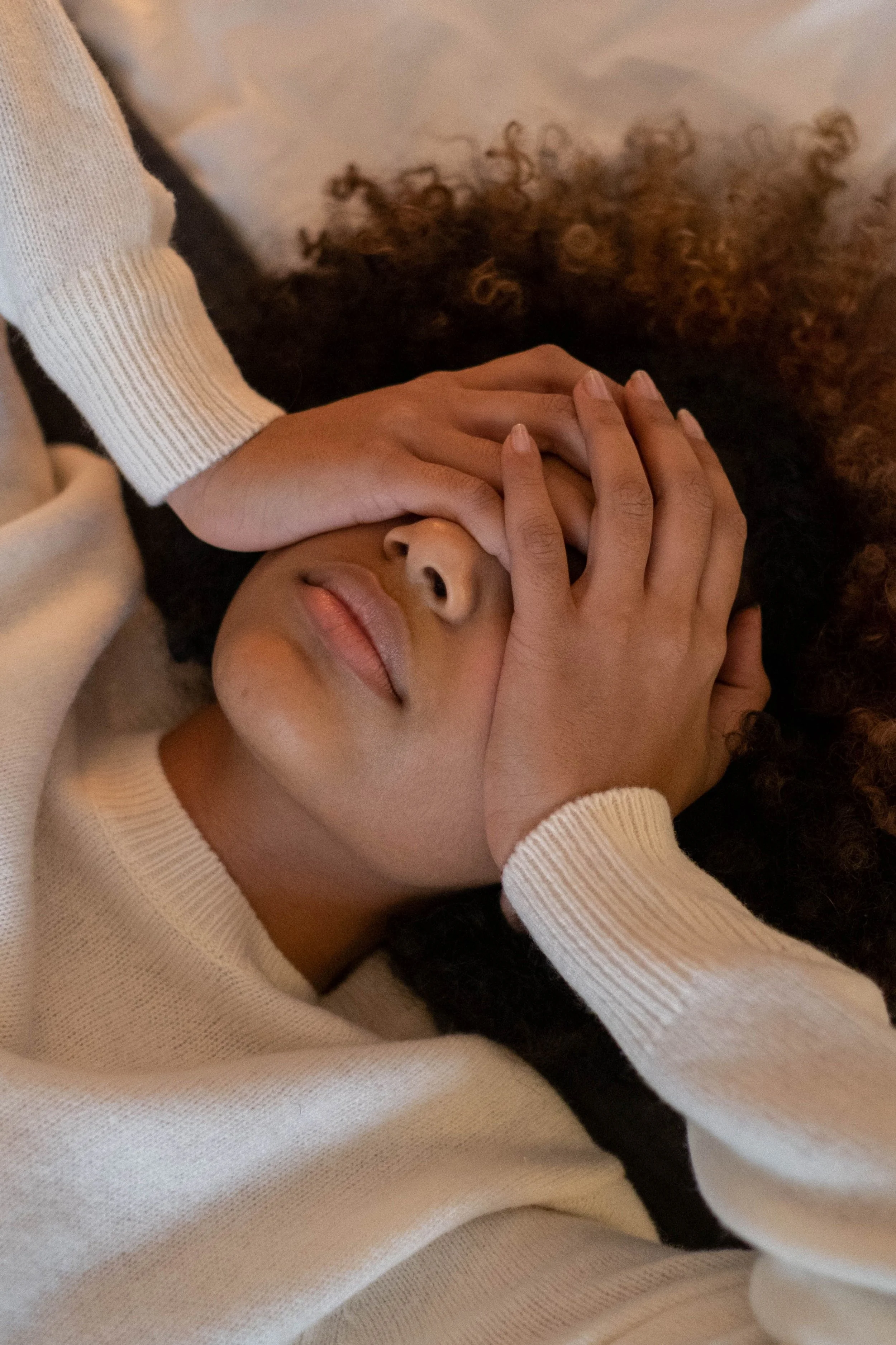 A woman lying down wearing a white long-sleeve shirt and resting her face on one hand. She has curly hair and appears calm.