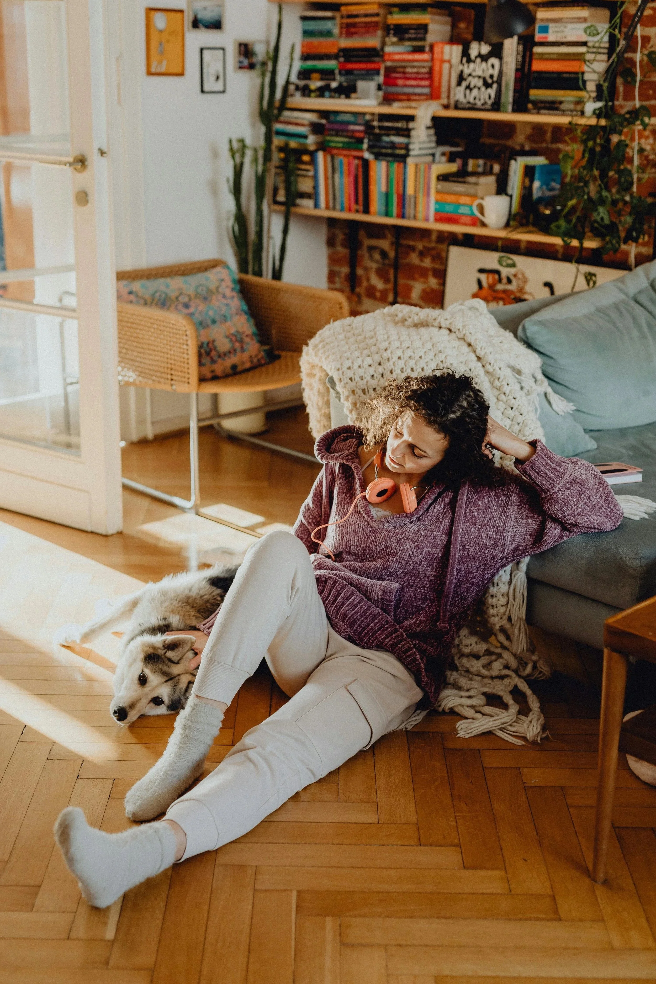 A woman with curly hair, wearing a purple hoodie and white pants, is sitting on a wooden floor, leaning against a couch, with a Siberian Husky puppy lying beside her. The woman appears relaxed, with one hand behind her head and the other near her face. The room is cozy, with a bookshelf filled with books and decorations, a wicker chair with a colorful cushion, and various plants.