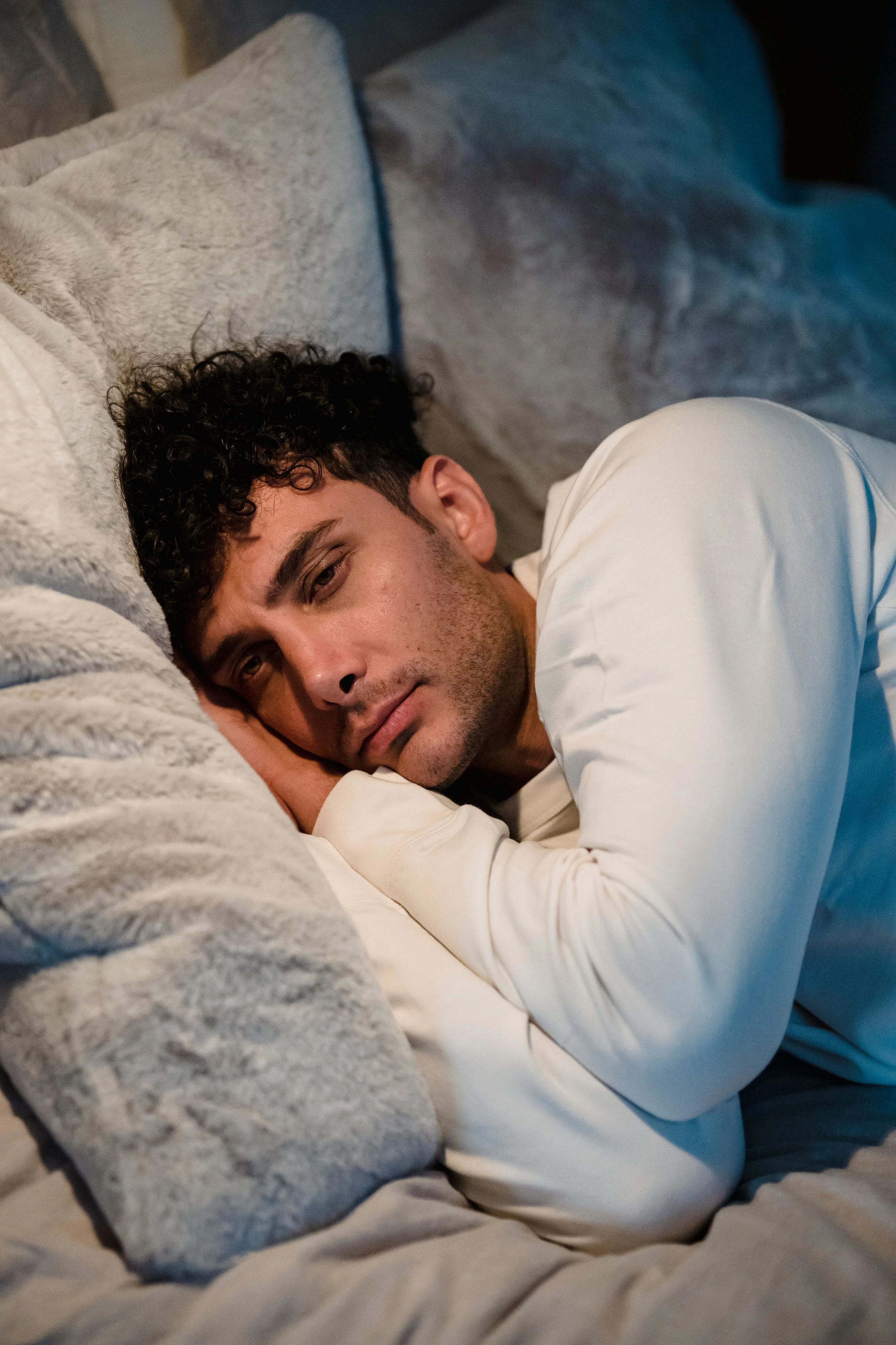 A young man with curly dark hair and light skin lying on a bed, resting his head on a fluffy gray pillow and looking at the camera.