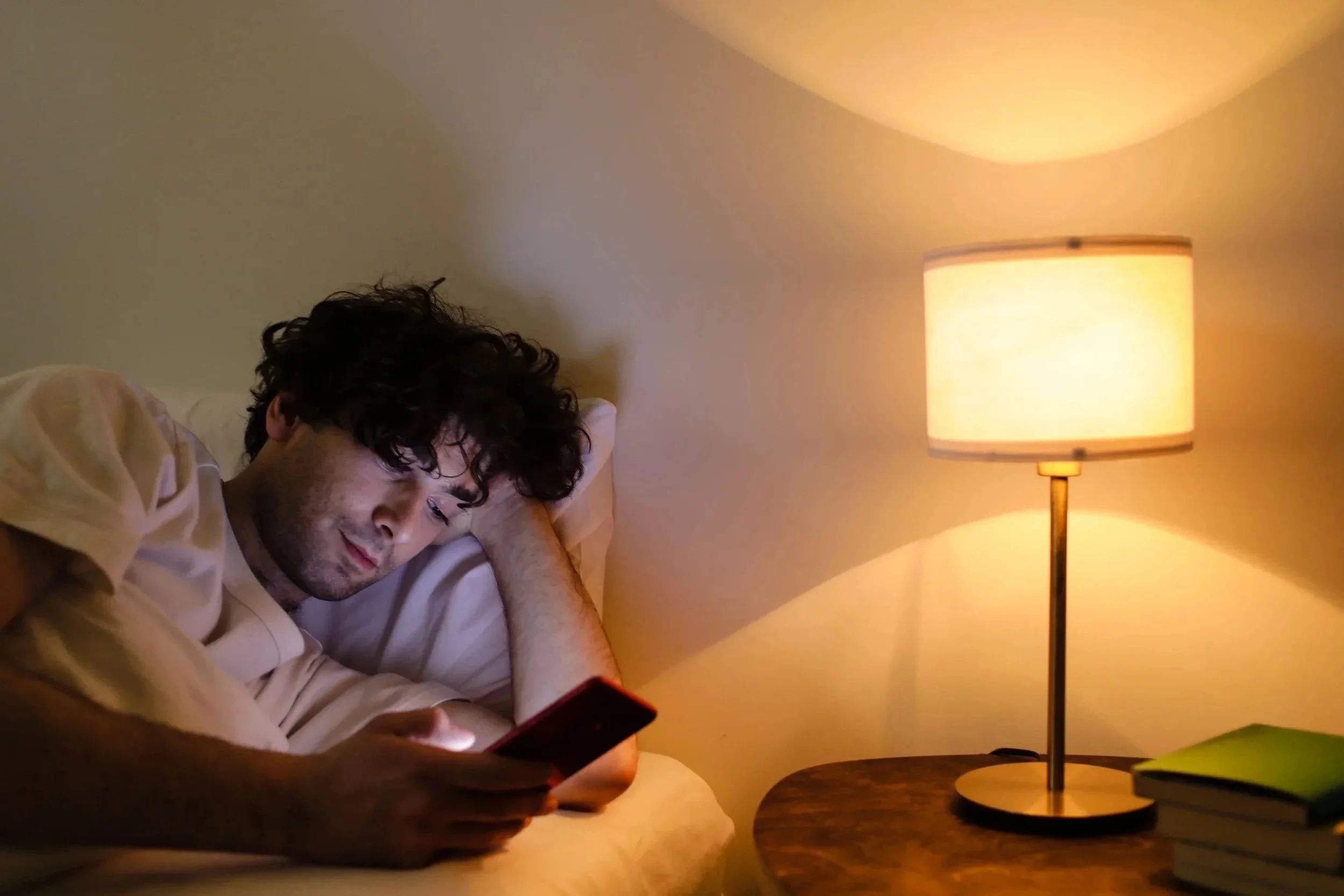 A young man with curly dark hair is lying in bed, looking at his phone. A warm-colored table lamp is on a nearby nightstand, creating a cozy atmosphere.