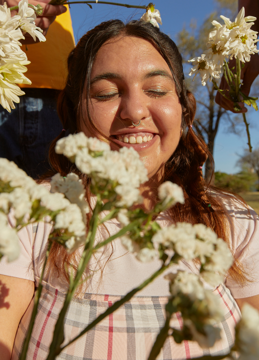 A woman with closed eyes smiling, surrounded by white flowers, outdoors on a sunny day.