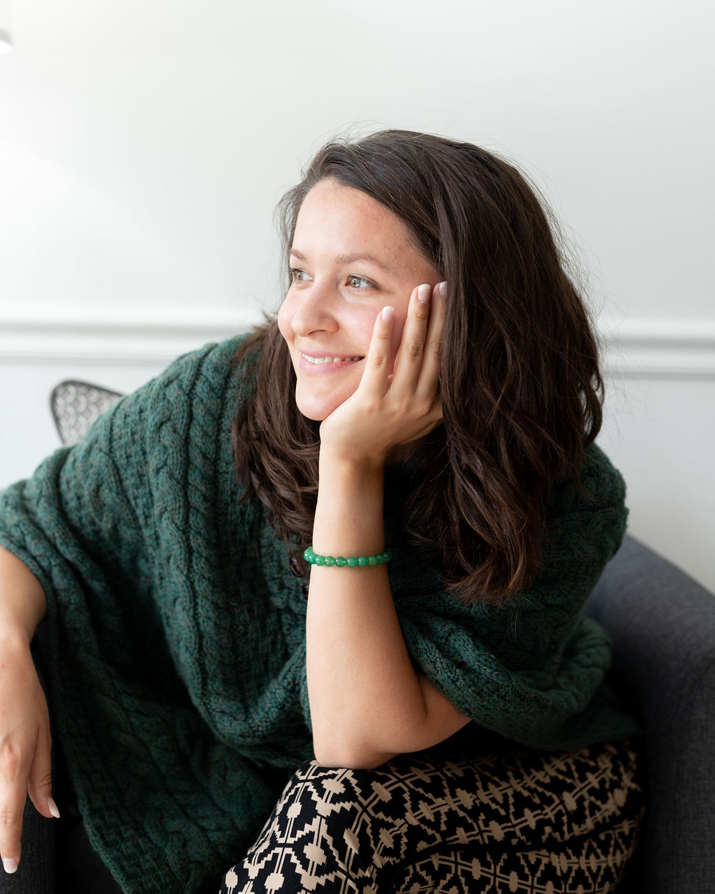A woman with brown hair, smiling and resting her face on her hand, wearing a green cable knit sweater and a green beaded bracelet, sitting on a dark gray couch in a bright room.