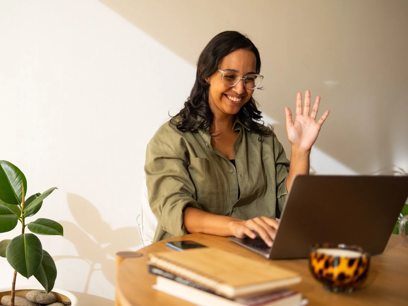 A woman with glasses and dark wavy hair smiling and waving while working on a laptop at a desk with notebooks, a phone, a cup, and a plant nearby.