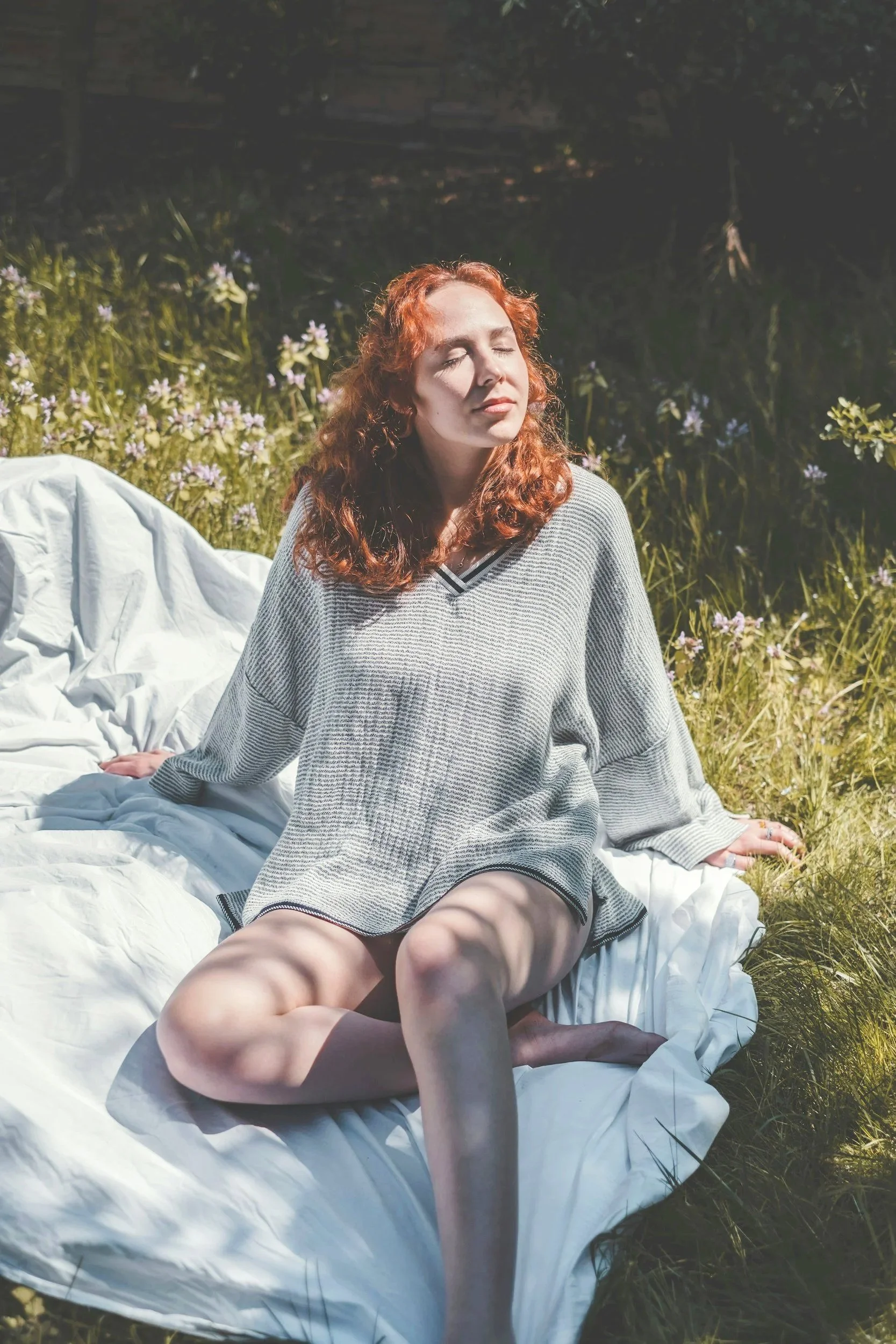 A woman with curly red hair sitting on a white blanket outdoors in a grassy area with small purple flowers, eyes closed, enjoying the sunlight.