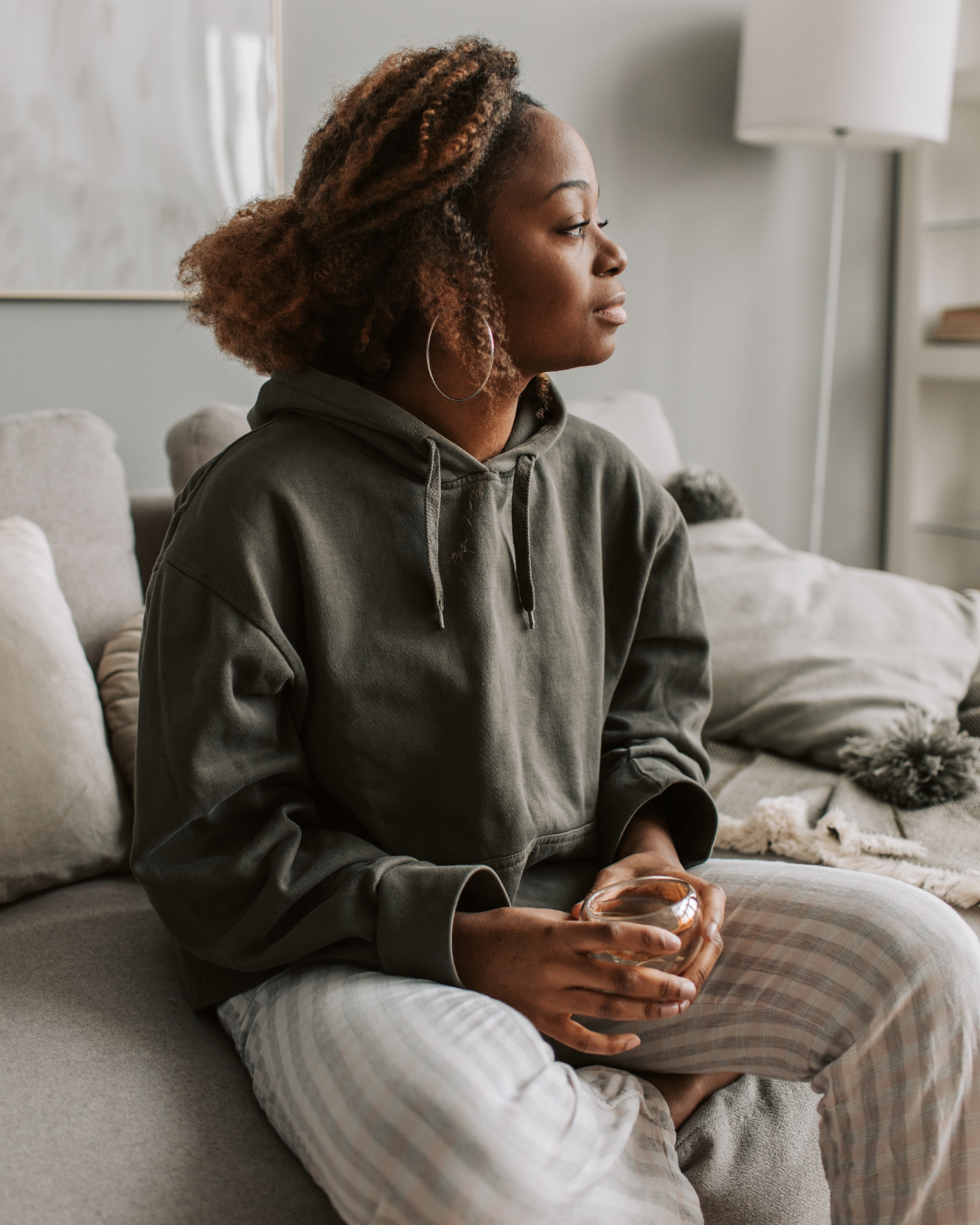 A woman with curly hair and hoop earrings sitting on a beige couch, wearing a gray hoodie and plaid pajama pants, holding a glass bowl in her hands, in a living room setting.