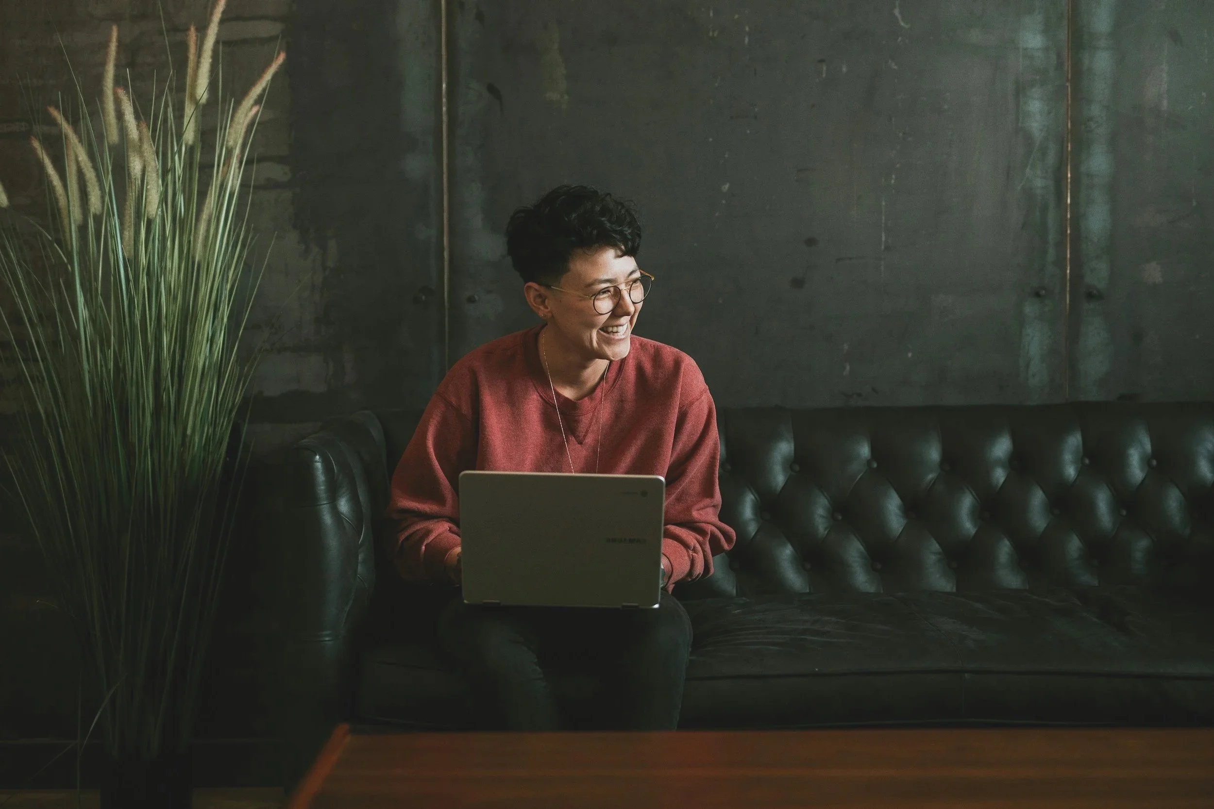 A woman with short dark curly hair, glasses, and a red sweater sitting on a black leather couch with a laptop, smiling and looking to her left, with green plants in the background.