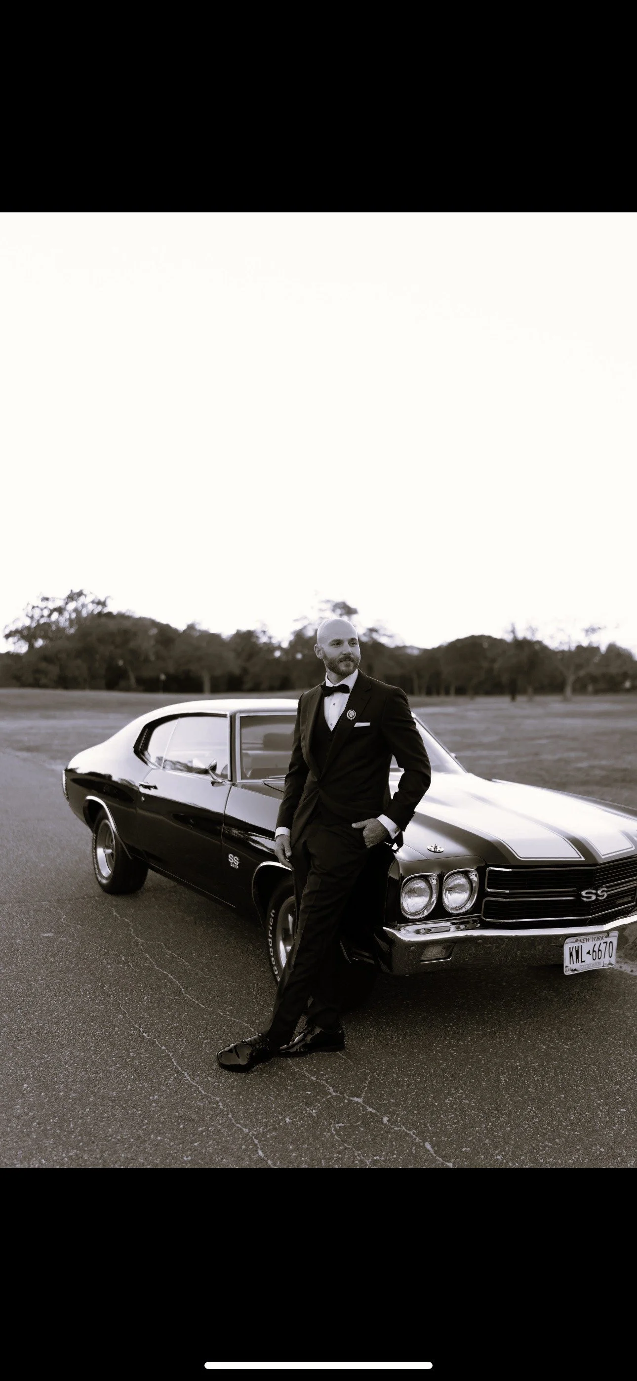 A man in a tuxedo stands next to a classic black Chevrolet Chevelle SS car on an empty road with trees in the background, black and white photo.