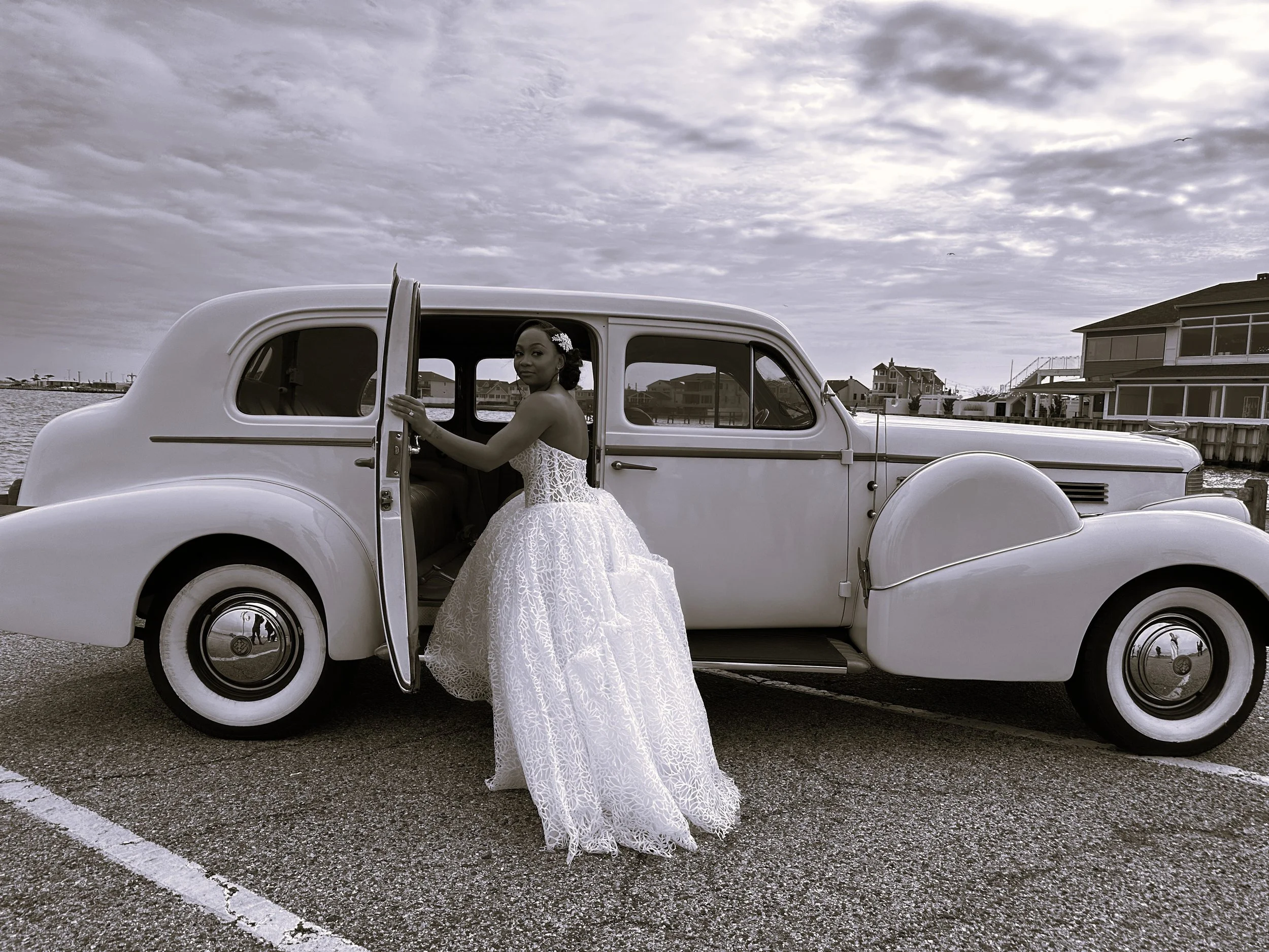 A woman in a white lace wedding dress standing next to an old fashioned white vintage car with the door open, near a waterfront with houses in the background, under cloudy sky.