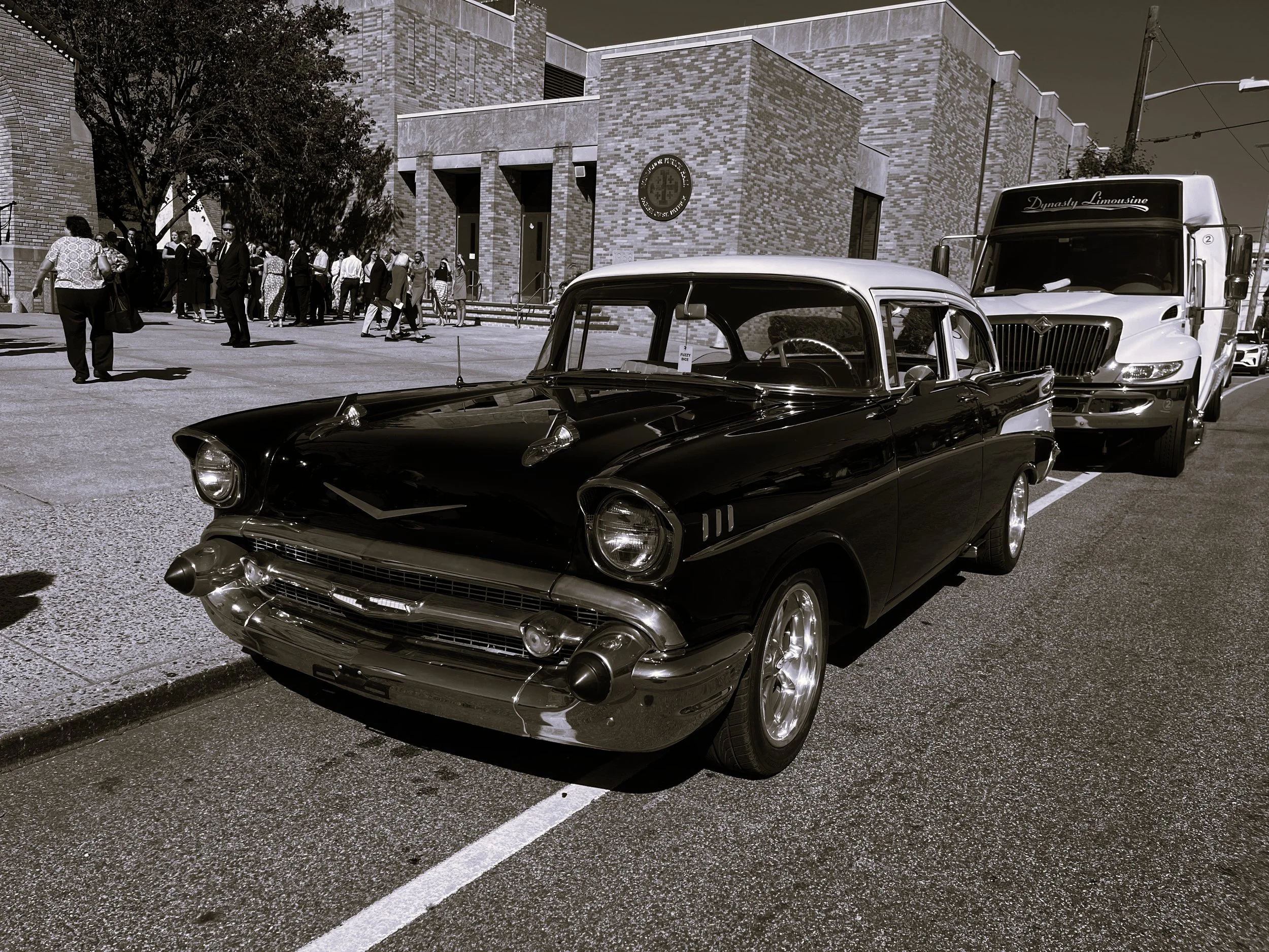A black vintage car parked on the street in front of a modern building with a group of people gathered in the background. There is also a large truck behind the car.