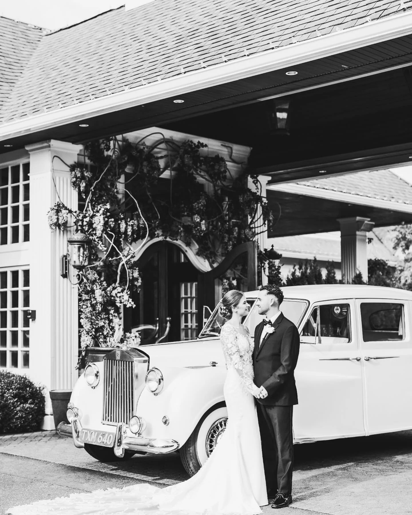 Black and white photo of a bride and groom standing in front of a vintage car at their wedding. The bride is wearing a long lace wedding dress, and the groom is dressed in a suit with a bow tie. They are holding hands and looking at each other, with a decorated building entrance in the background.