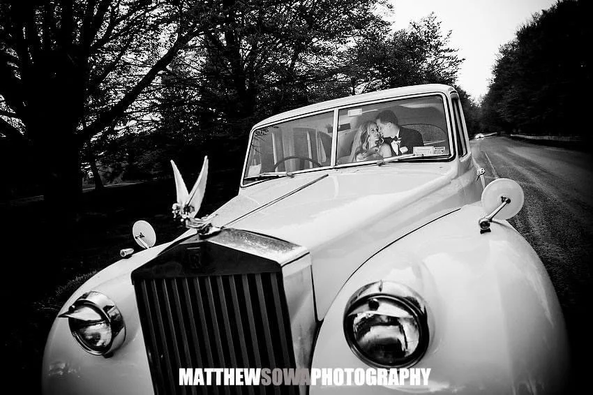 A vintage white car with two people inside, in a rural setting with trees on either side of the road. The couple appears to be looking at each other through the windshield.