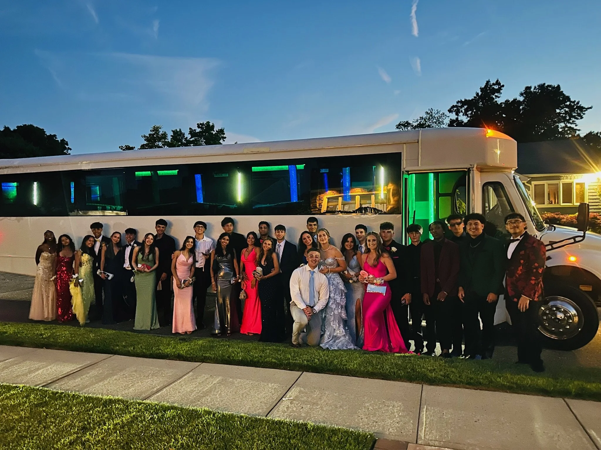 A large group of young people in formal attire posing in front of a white party bus during dusk. The group includes males and females dressed in suits, dresses, and gowns, with some holding small bags or clutch purses. The party bus has green and blue lighting inside and is parked on a street with a house visible in the background.