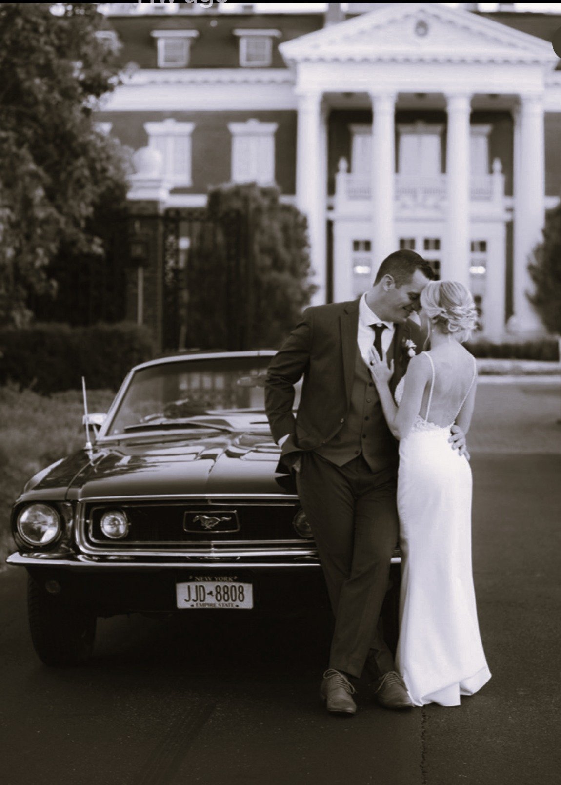A couple in wedding attire leaning on a black vintage Ford Mustang in front of a grand, classical-style building with columns.