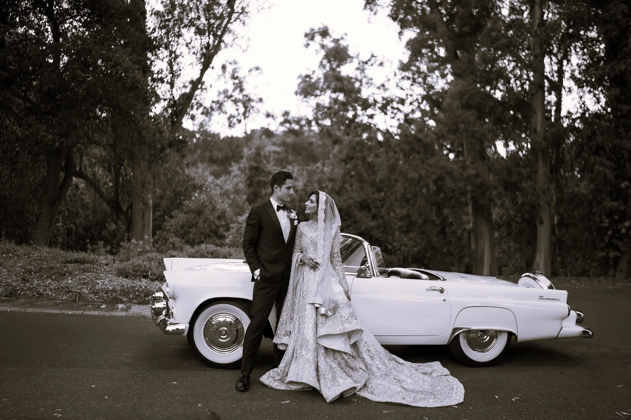 A newlywed couple in wedding attire standing in front of a vintage car on a park road with trees in the background, black and white photo.