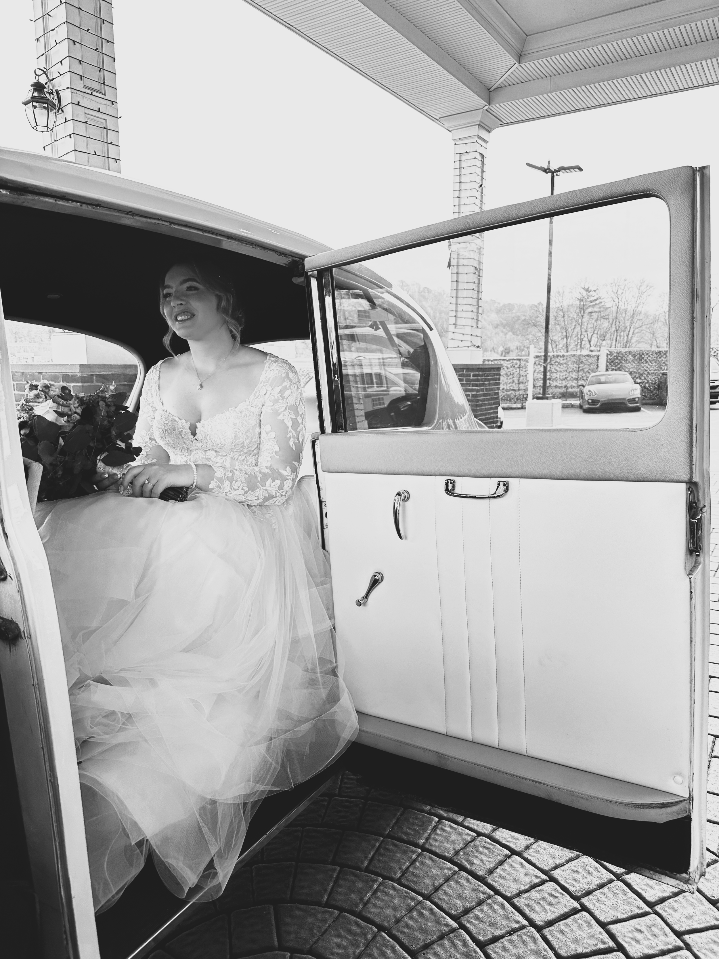 A bride sitting inside a vintage car with the door open, wearing a lace wedding dress and holding a bouquet.