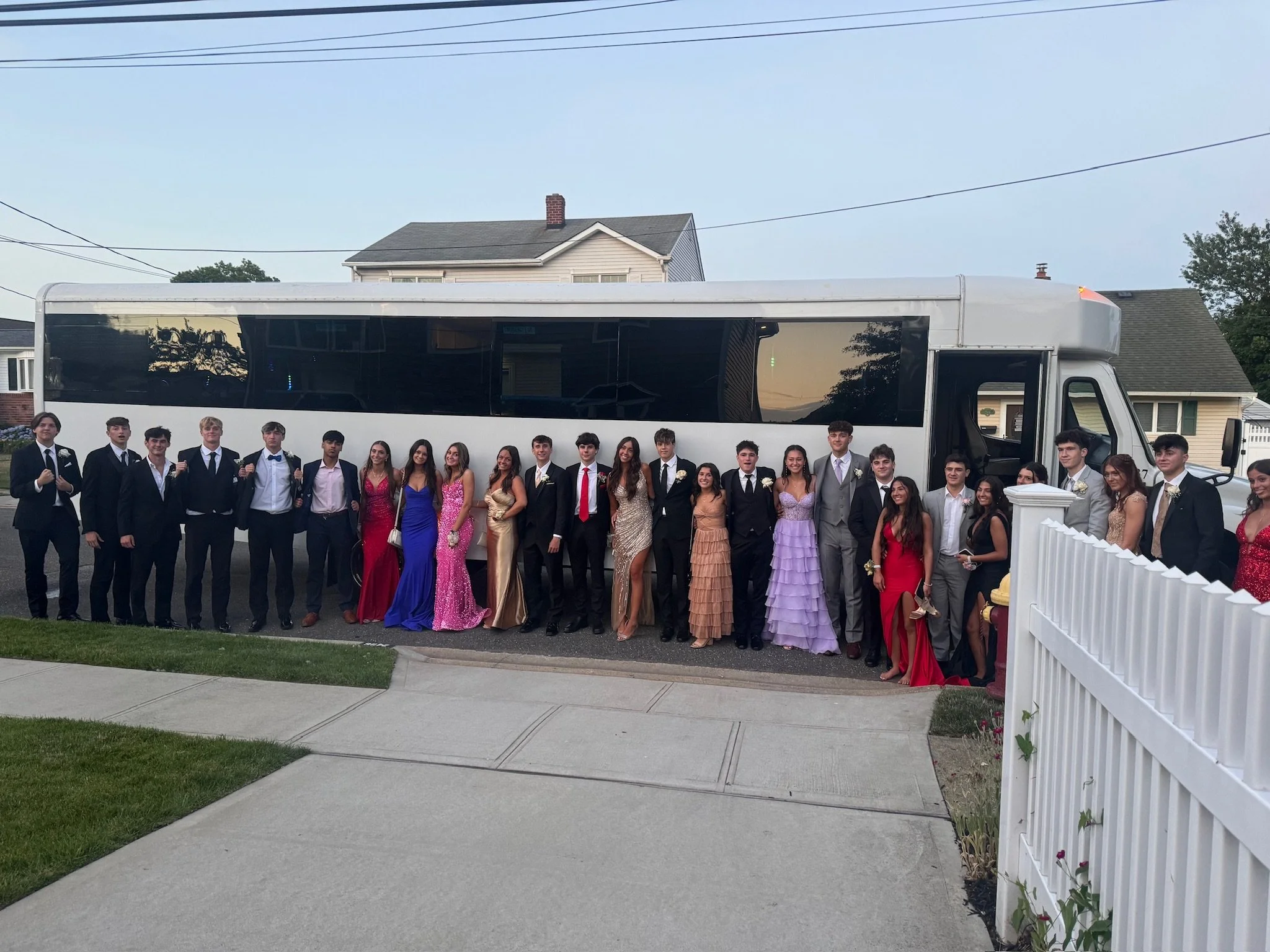 Group of young men and women dressed in formal attire, standing in front of a bus, during what appears to be a prom or formal event in a suburban neighborhood.