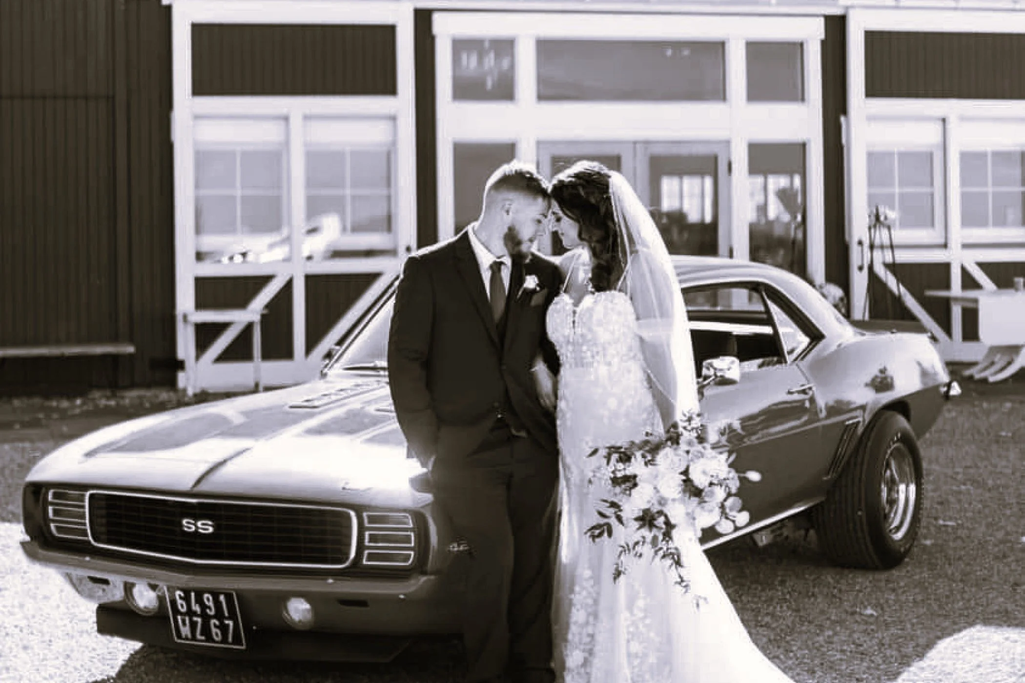 Black and white photo of a bride and groom leaning their foreheads together in front of a vintage black car, outside a building with large windows and a porch.