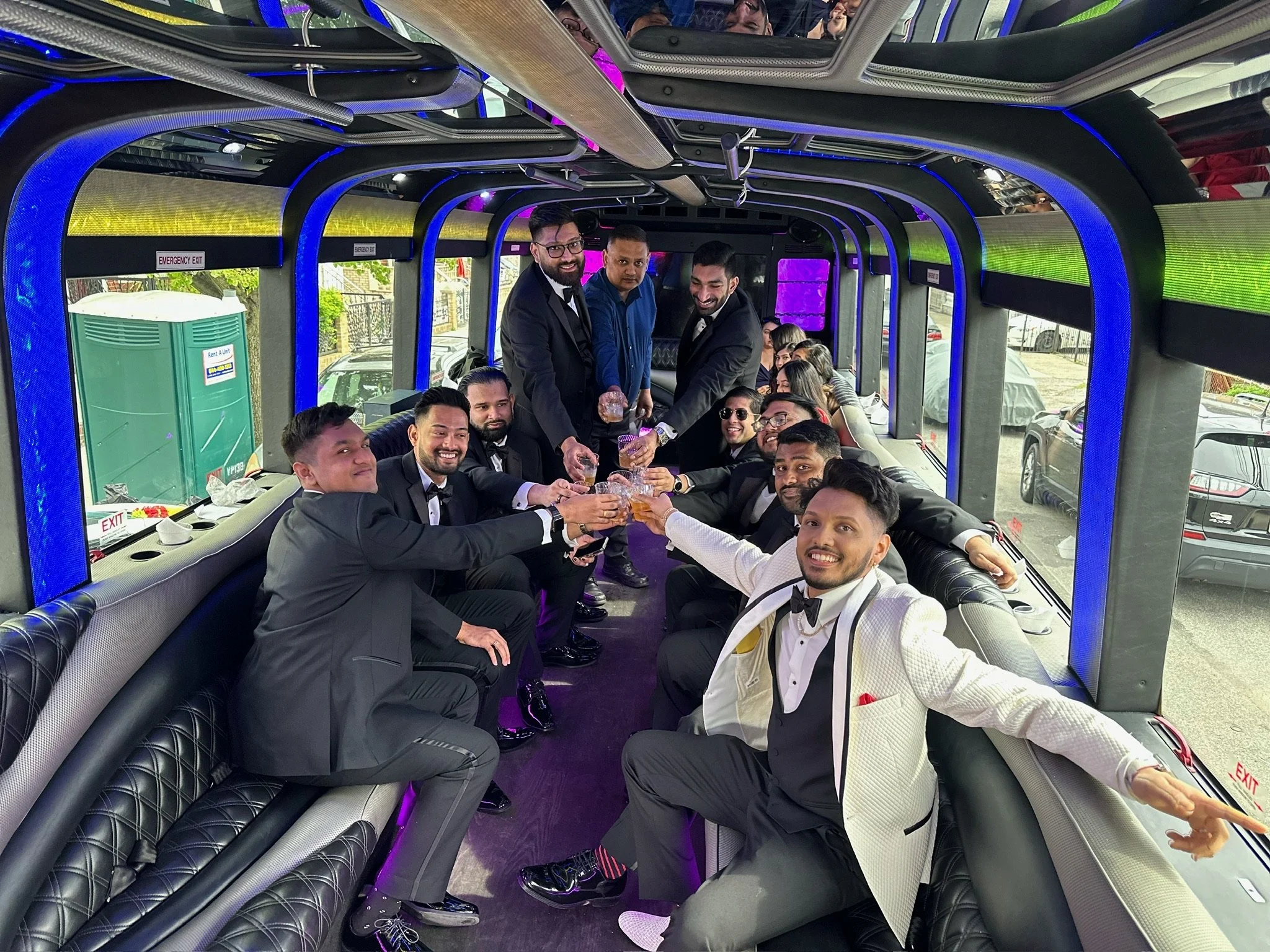 A group of men dressed in tuxedos and suits on a party bus, toasting with drinks and smiling for the camera.