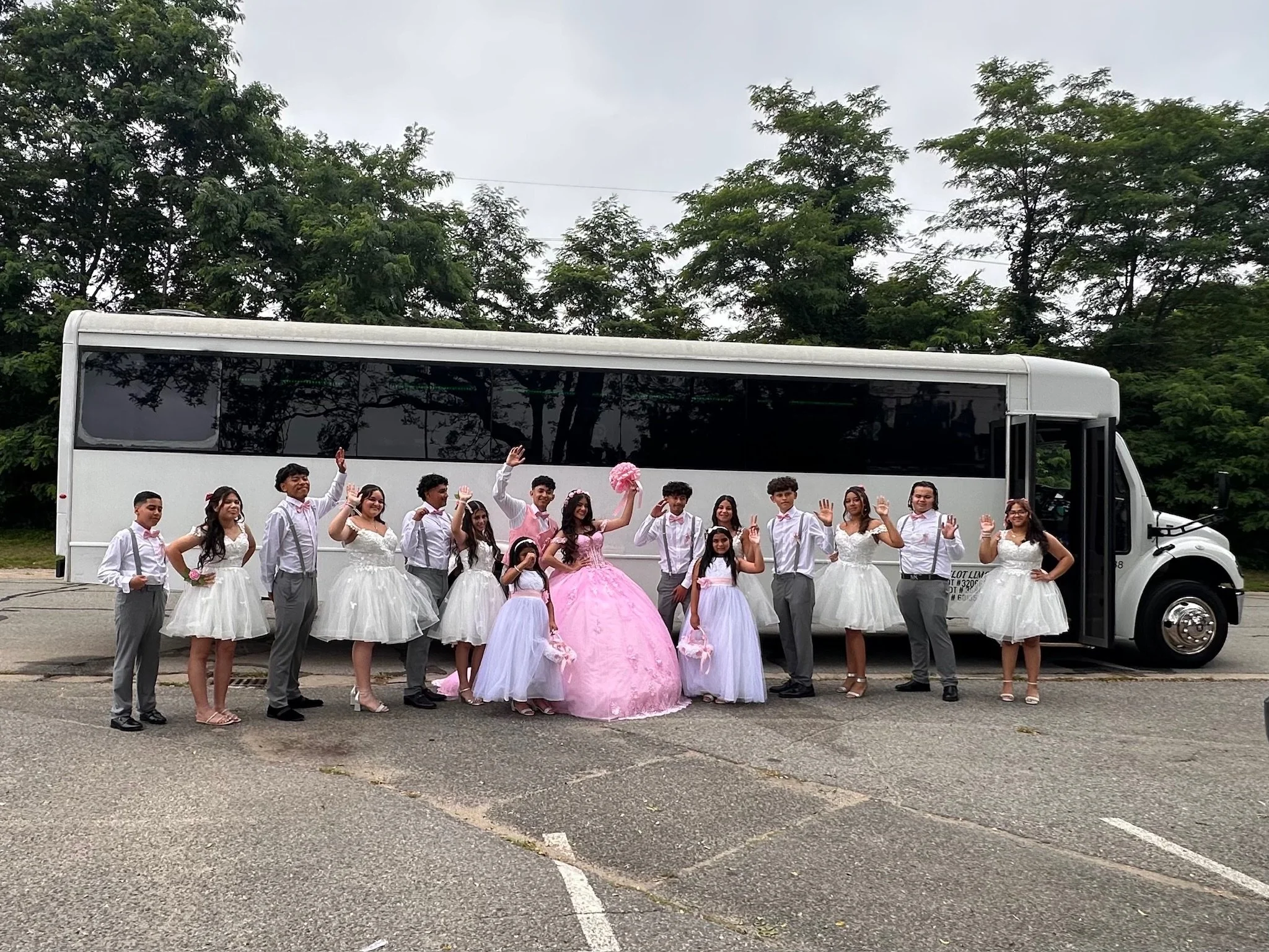 Group of people in formal dresses and suits standing in front of a bus, some waving, outdoors on a cloudy day.