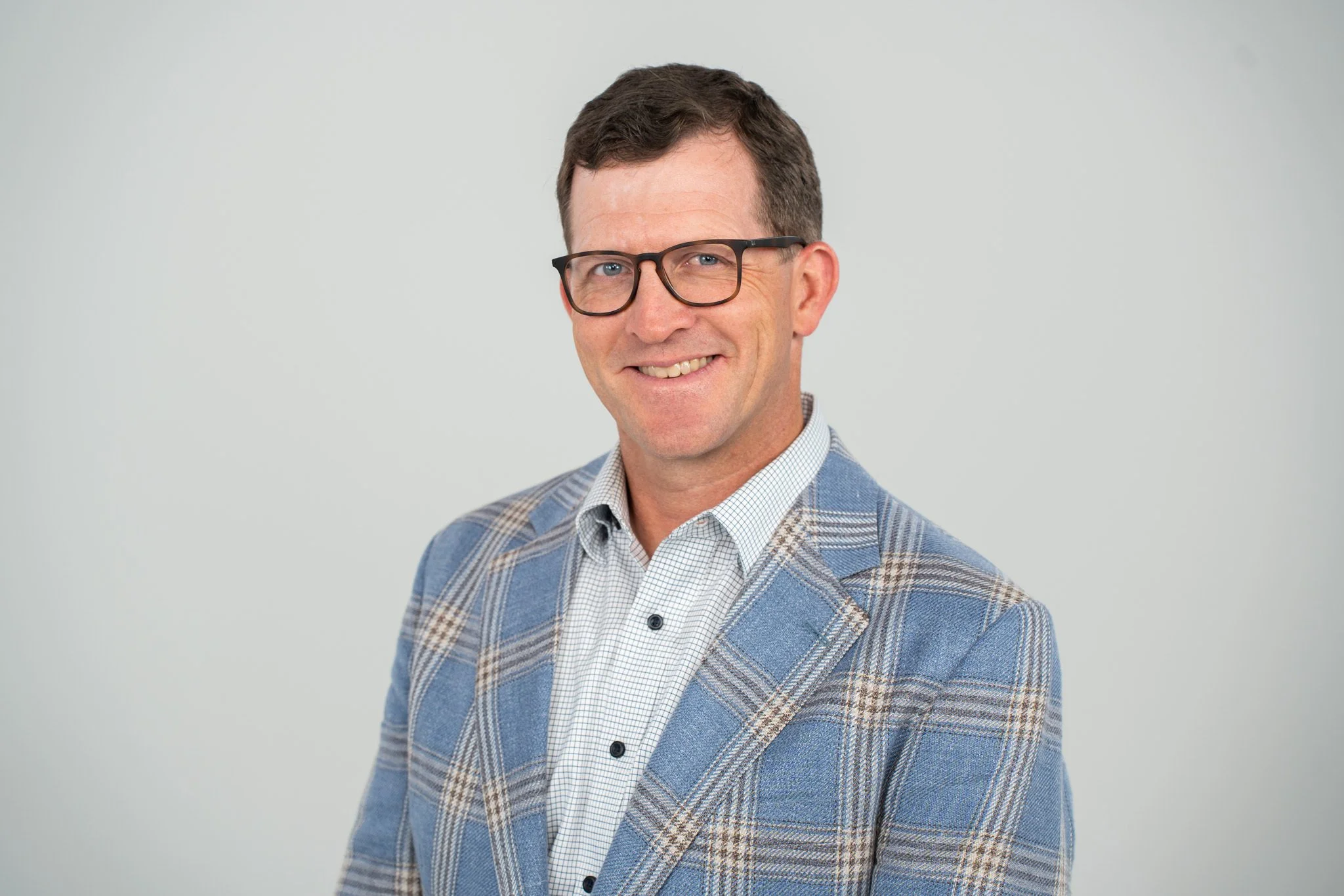 Headshot of a man wearing glasses, a navy blue suit, a white shirt, and a pink patterned tie, smiling against a gray background.