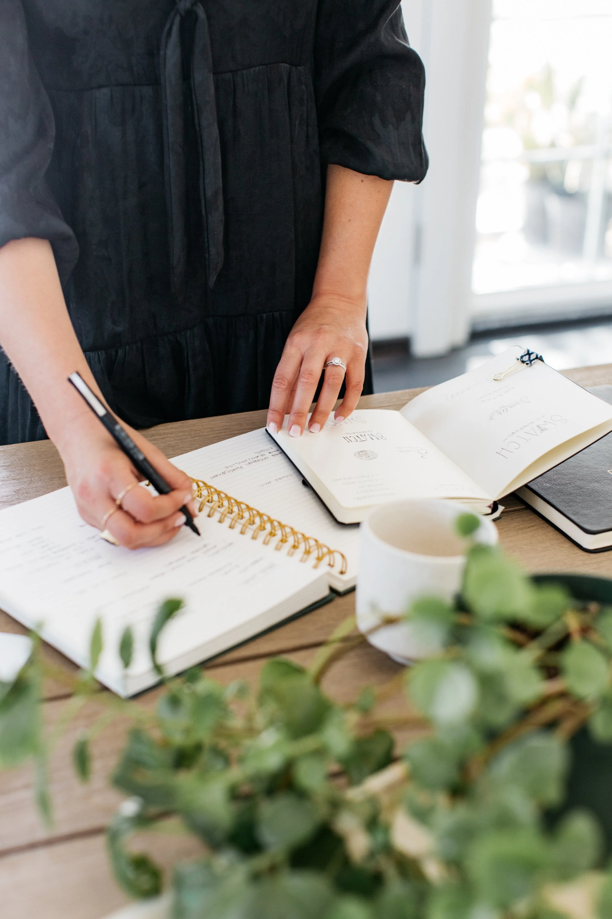 Person writing in a planner at a wooden table with open notebooks, a cup of coffee, and a plant in the foreground.