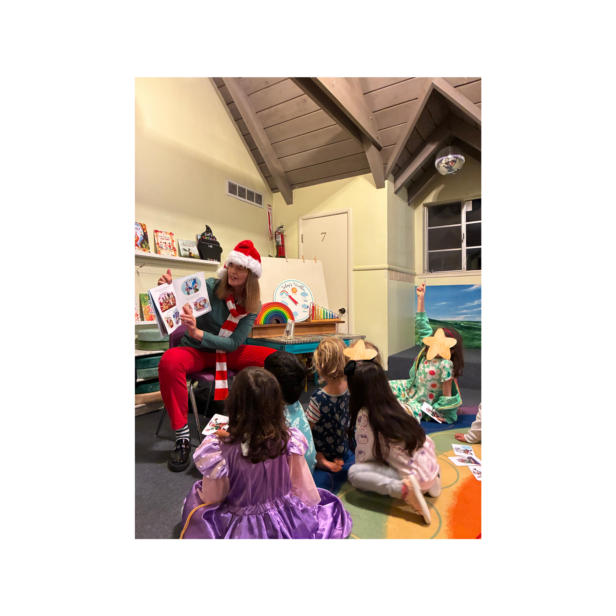 co-author Ellen H.S., wearing red pants, a striped scarf, and a Santa hat, reads a book to young children.